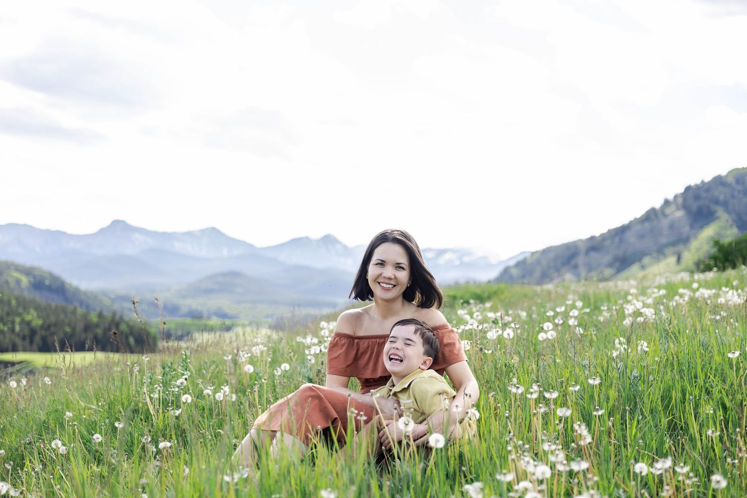 Mom and son sitting in a wildflower meadow with mountain views, smiling joyfully on a cloudy day.