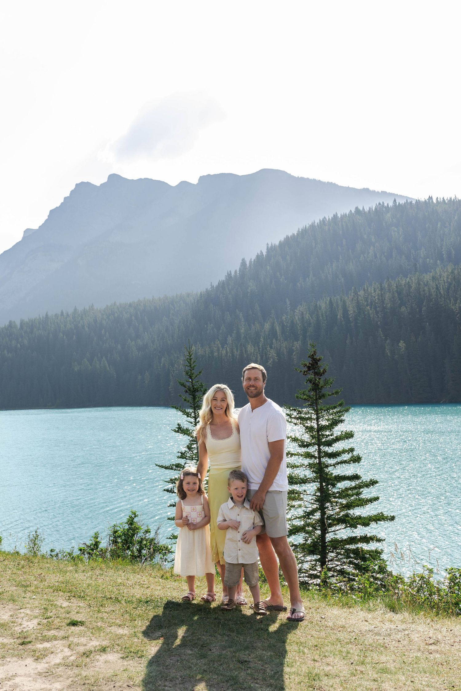 Family of four stands by a turquoise mountain Lake Minnewanka surrounded by pine trees and mountains.in Banff