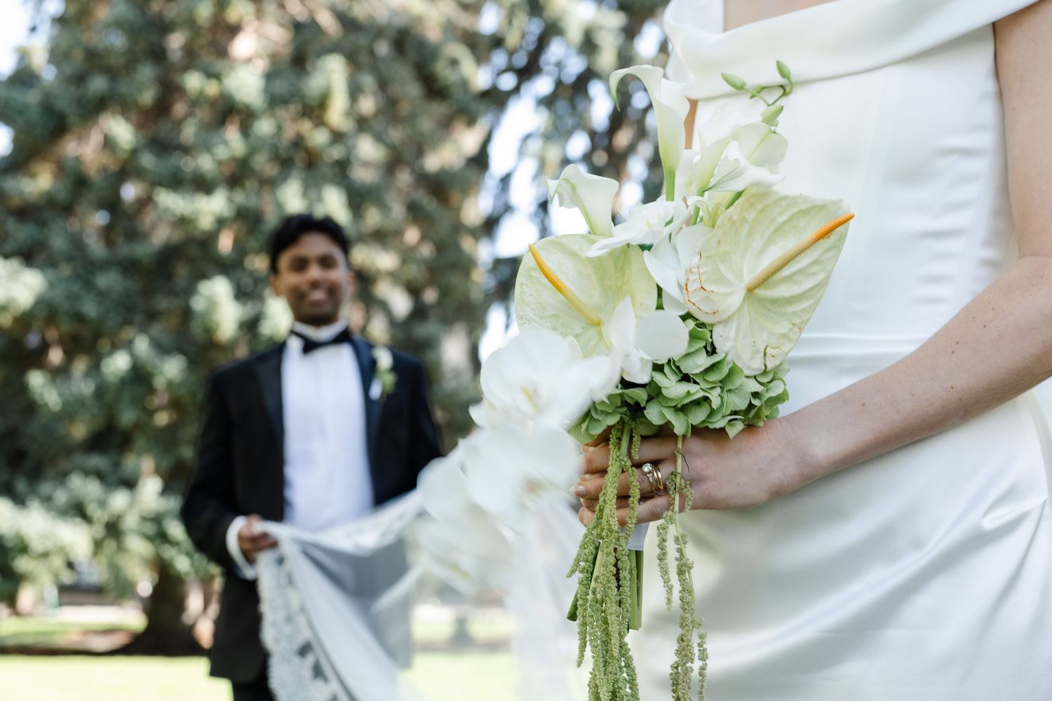 Bride holding white calla lily bouquet with greenery, groom in tuxedo standing in background at outdoor wedding.