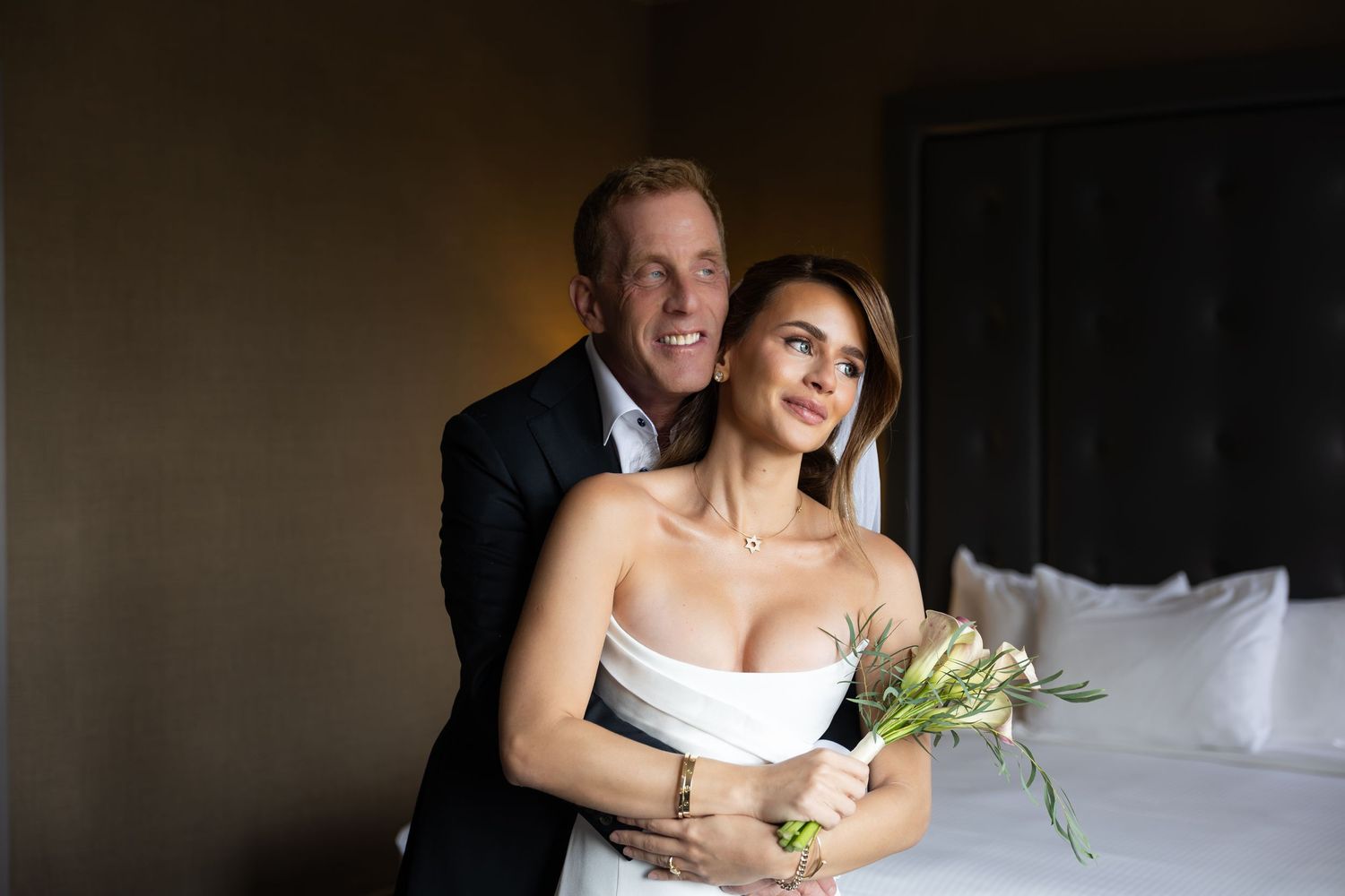 Couple embracing in a hotel room, woman in white strapless dress holding flowers, man in dark suit behind her.