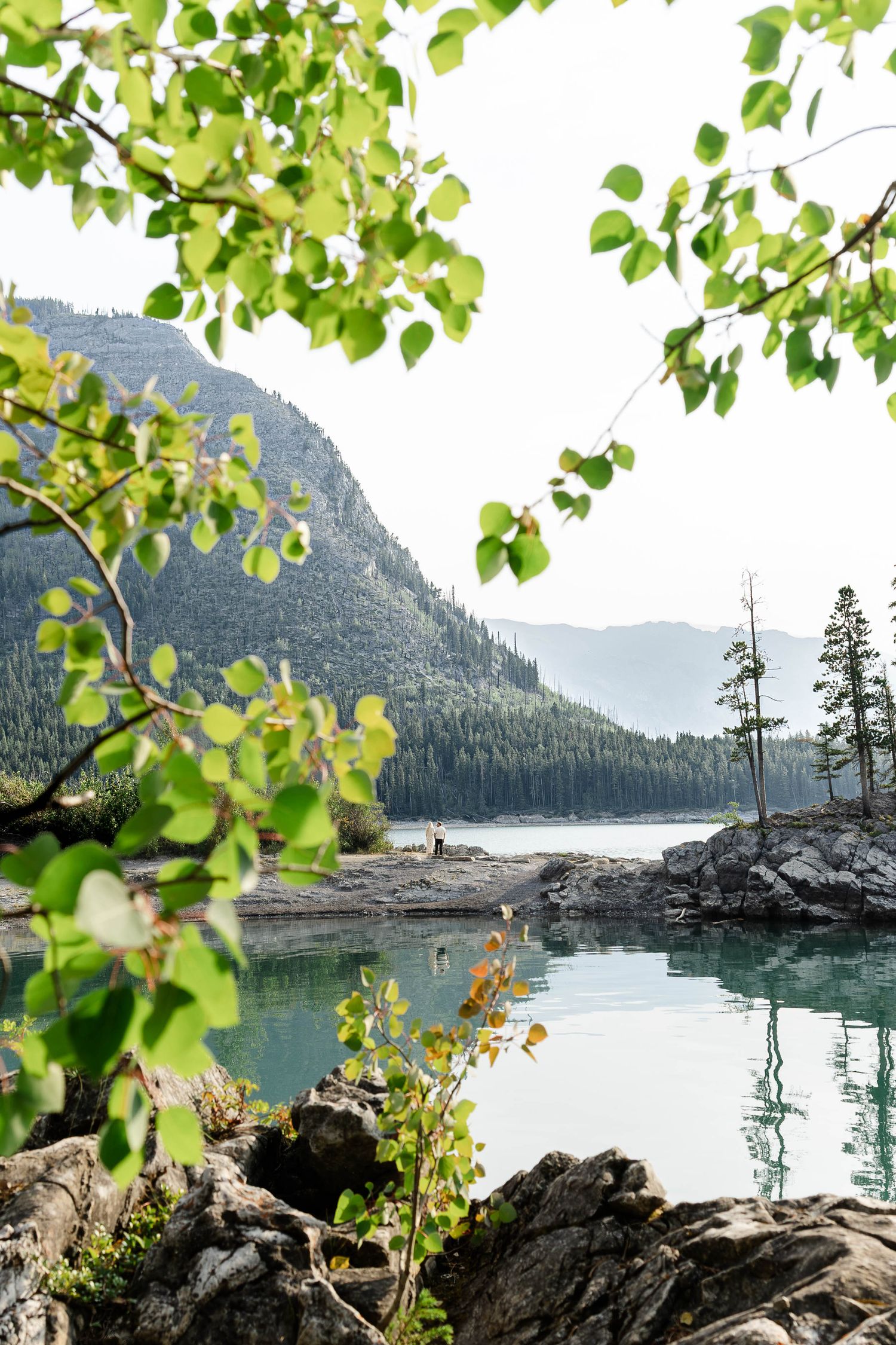 Serene mountain lake framed by green birch leaves with rocky shores, evergreen forest.Romantic couple in Minnewanka Lake in Banff.