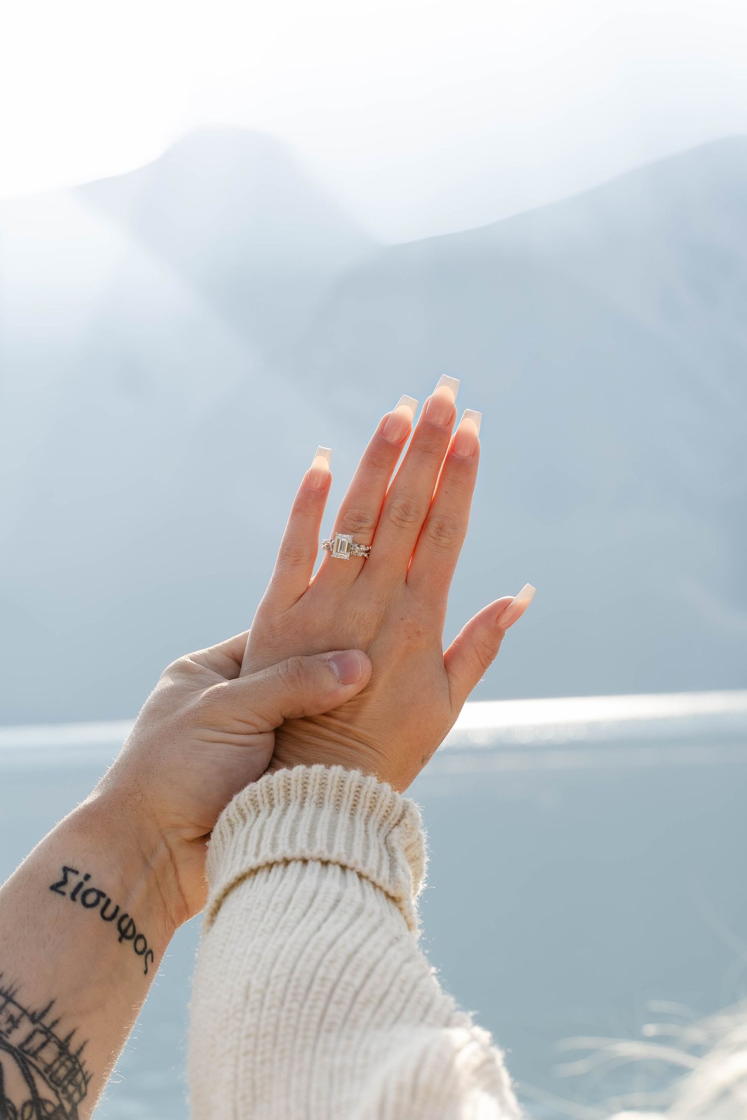 Two hands raised against mountain backdrop, woman showing diamond engagement ring, tattoo visible on man's wrist.Romantic couple in Minnewan