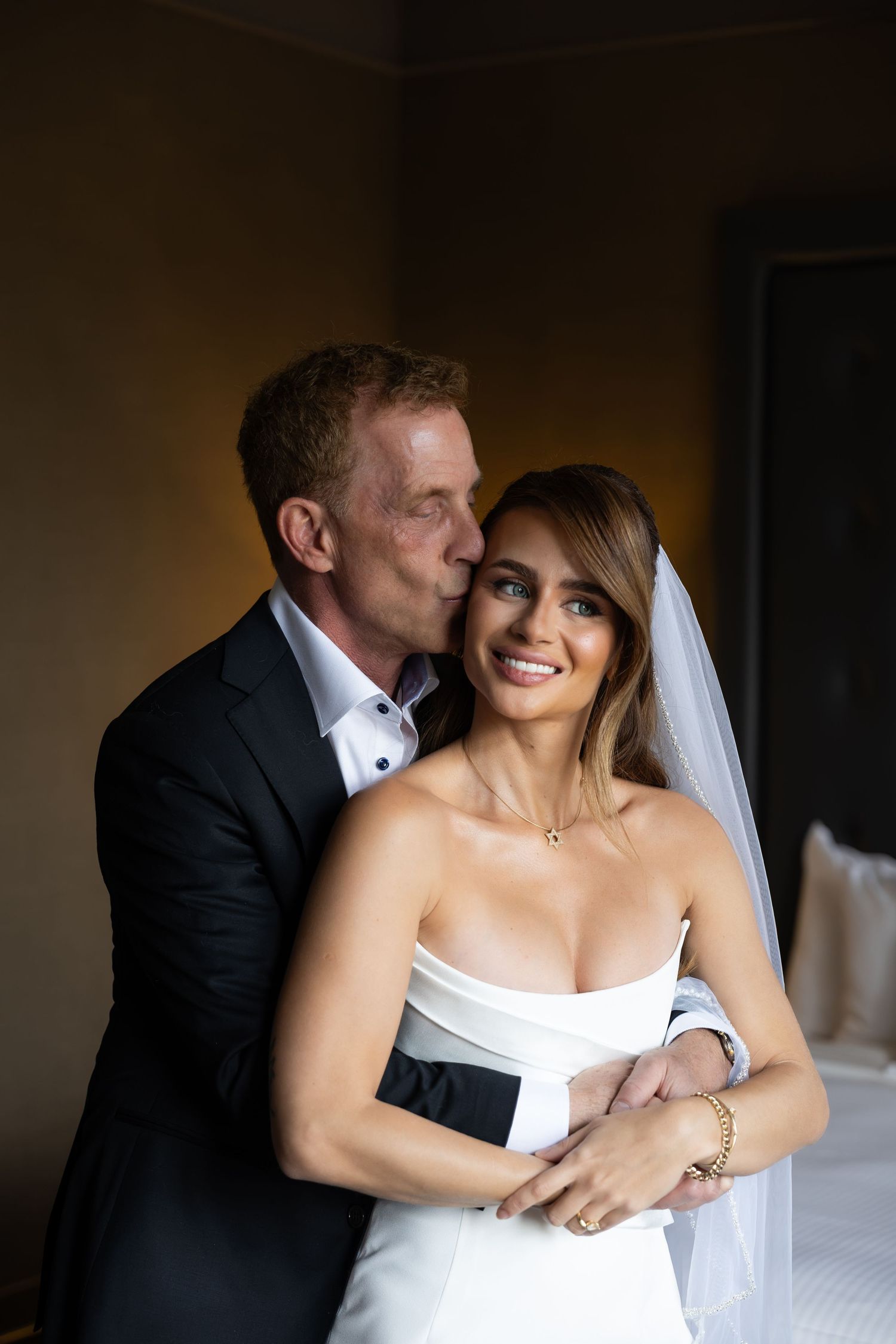 Groom in black suit kissing bride in white strapless gown and veil on the cheek while she smiles.