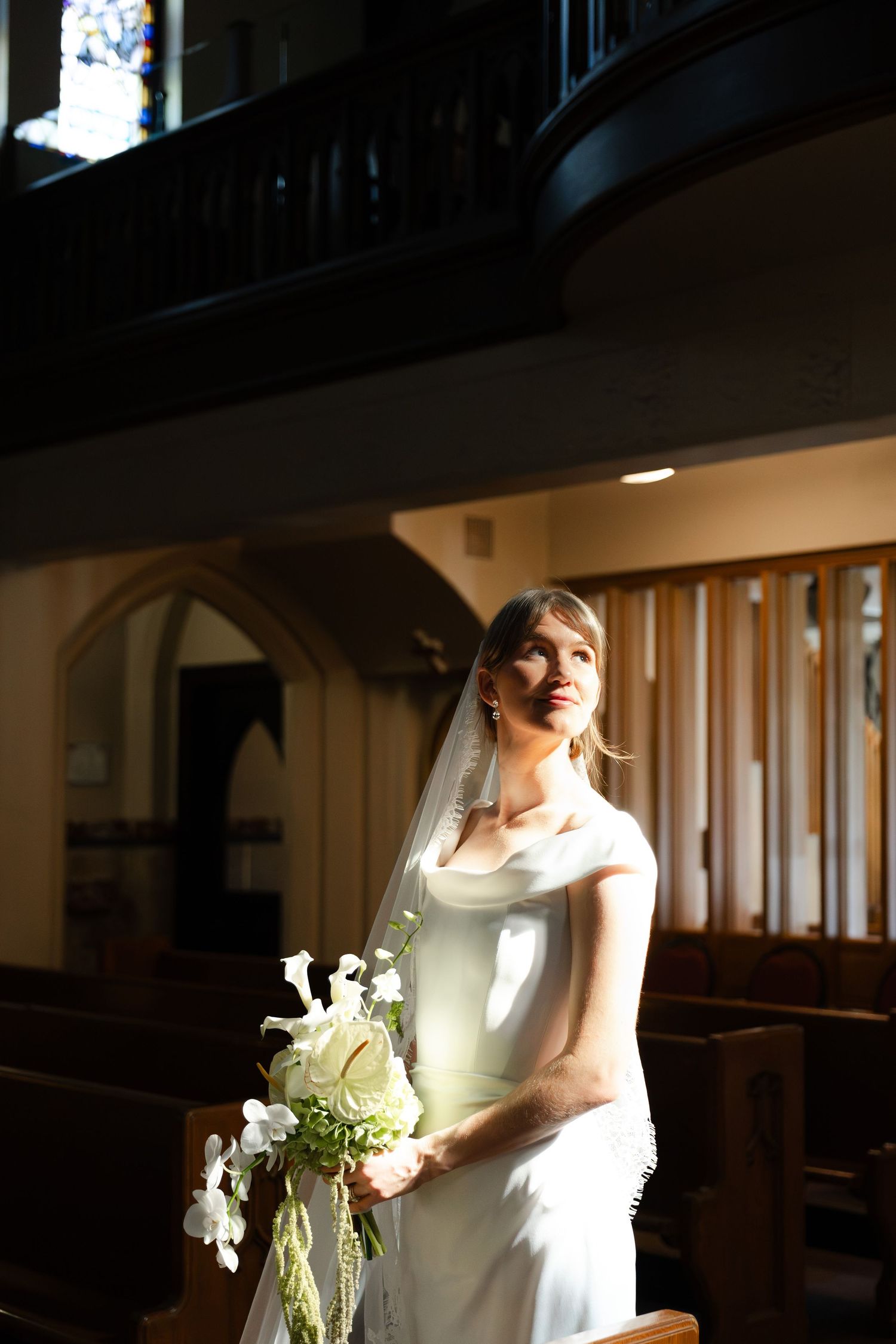 Bride in white gown and veil holds bouquet of white flowers inside a sunlit church.