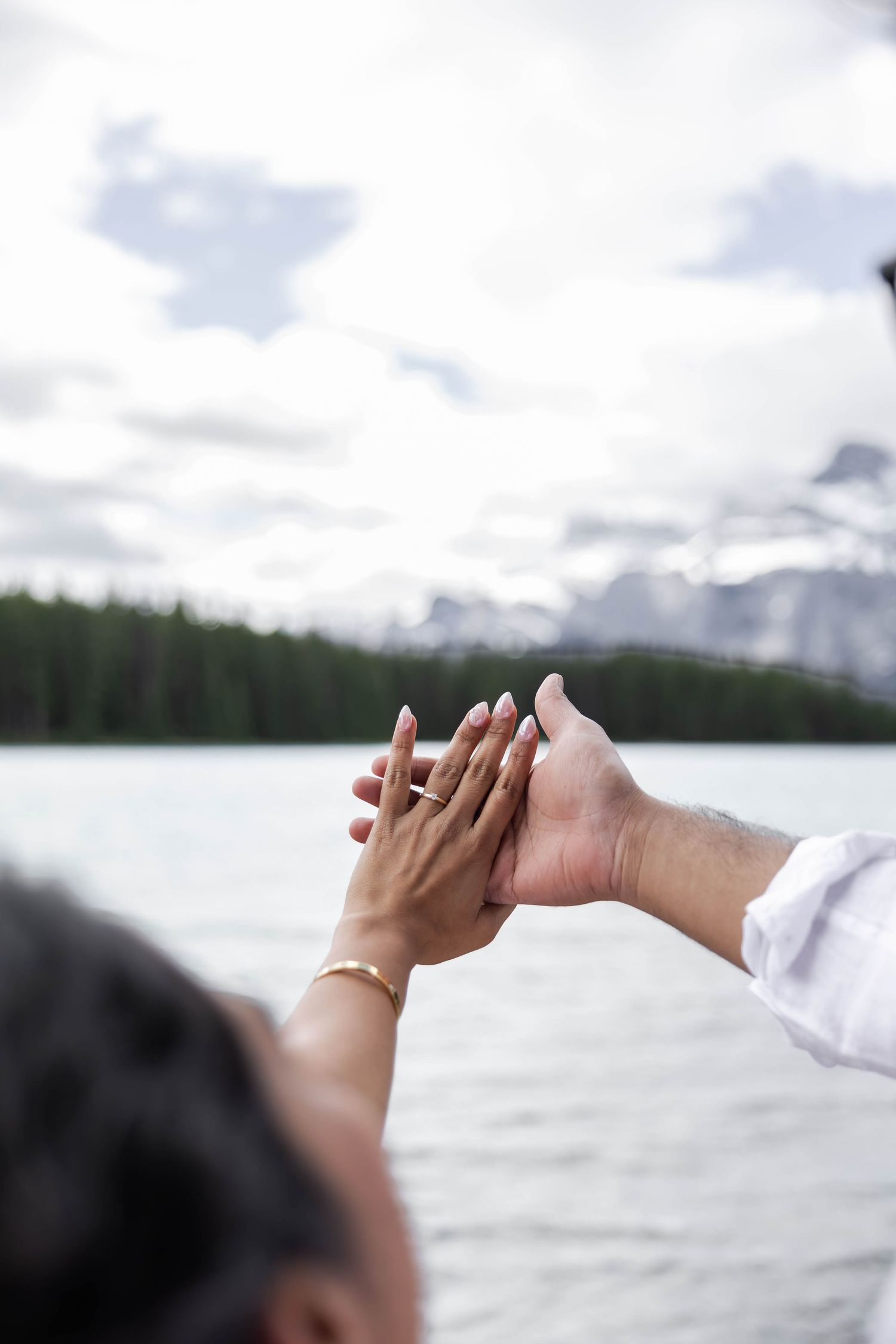 Couple holding hands together over a scenic mountain lake with forest and cloudy sky backdrop.