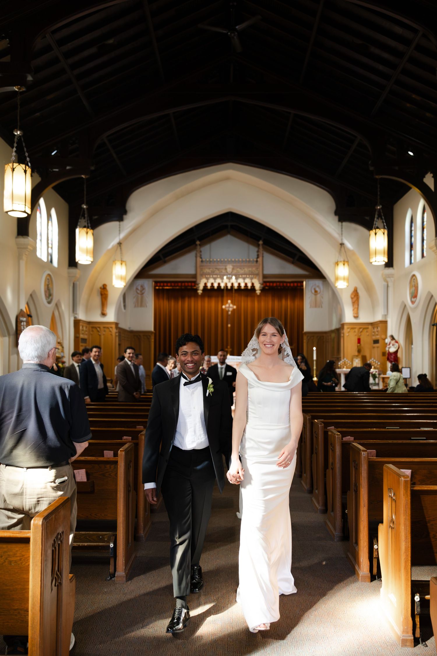 A joyful bride and groom walk hand in hand down a sunlit church aisle after their wedding.
