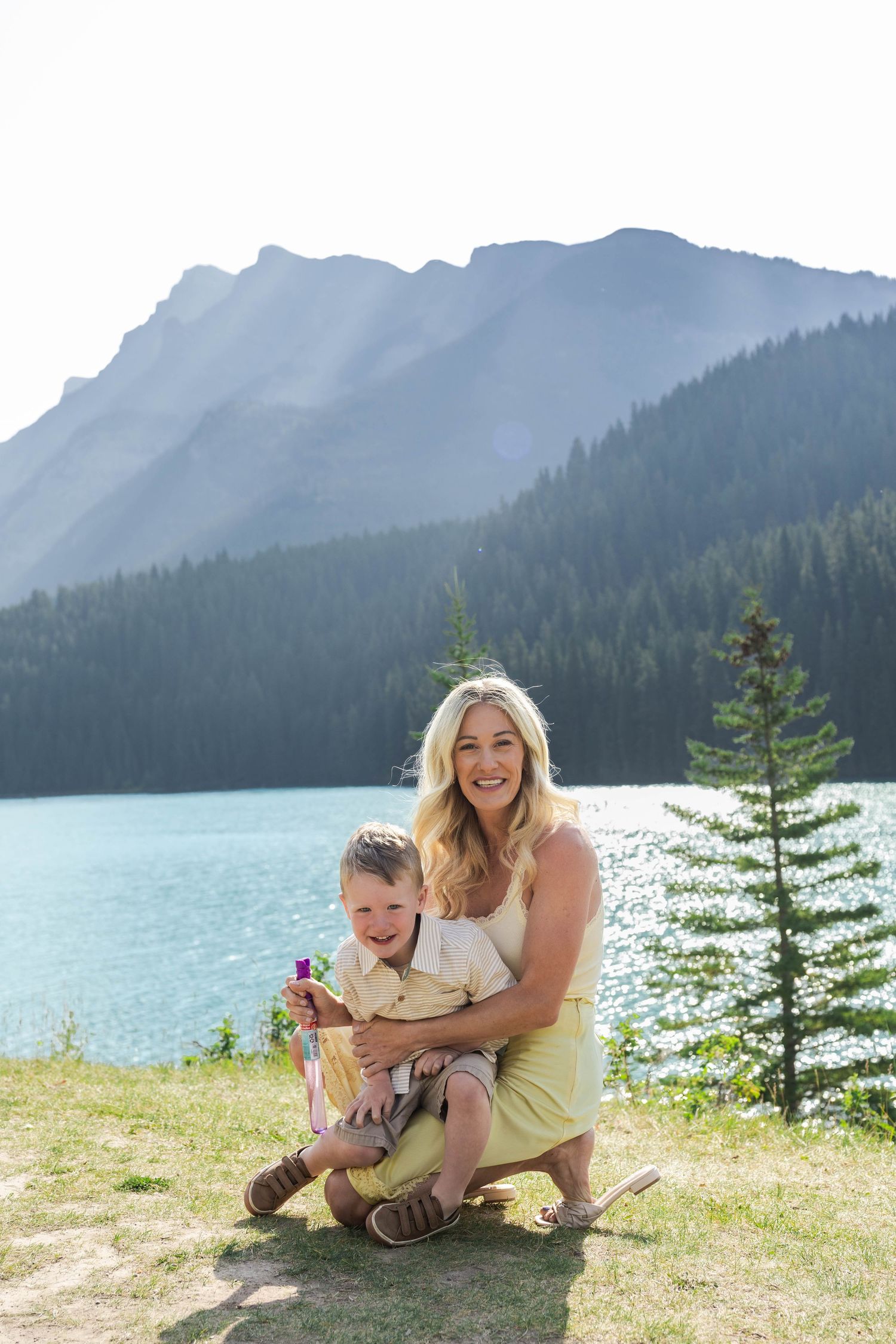 A smiling blonde woman crouches with a toddler by a turquoise mountain Lake Minnewanka surrounded by pine trees.in Banff