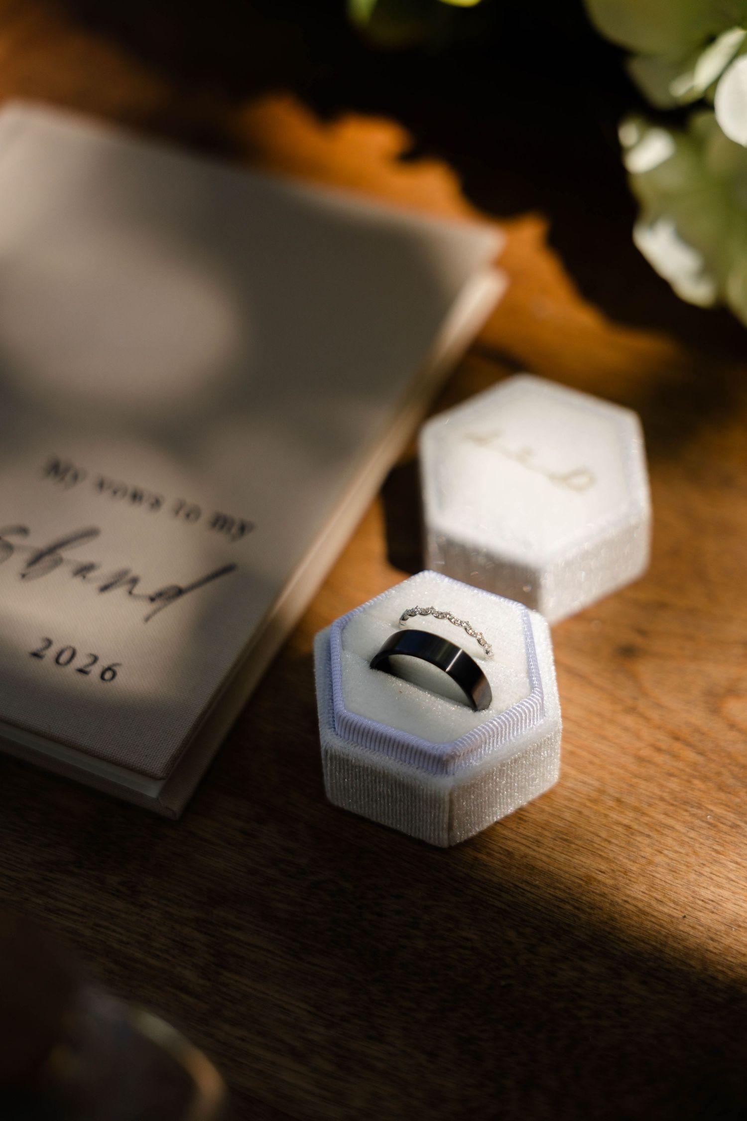 A silver wedding band in a white hexagonal velvet ring box beside a vows booklet dated 2026 on a wooden surface.
