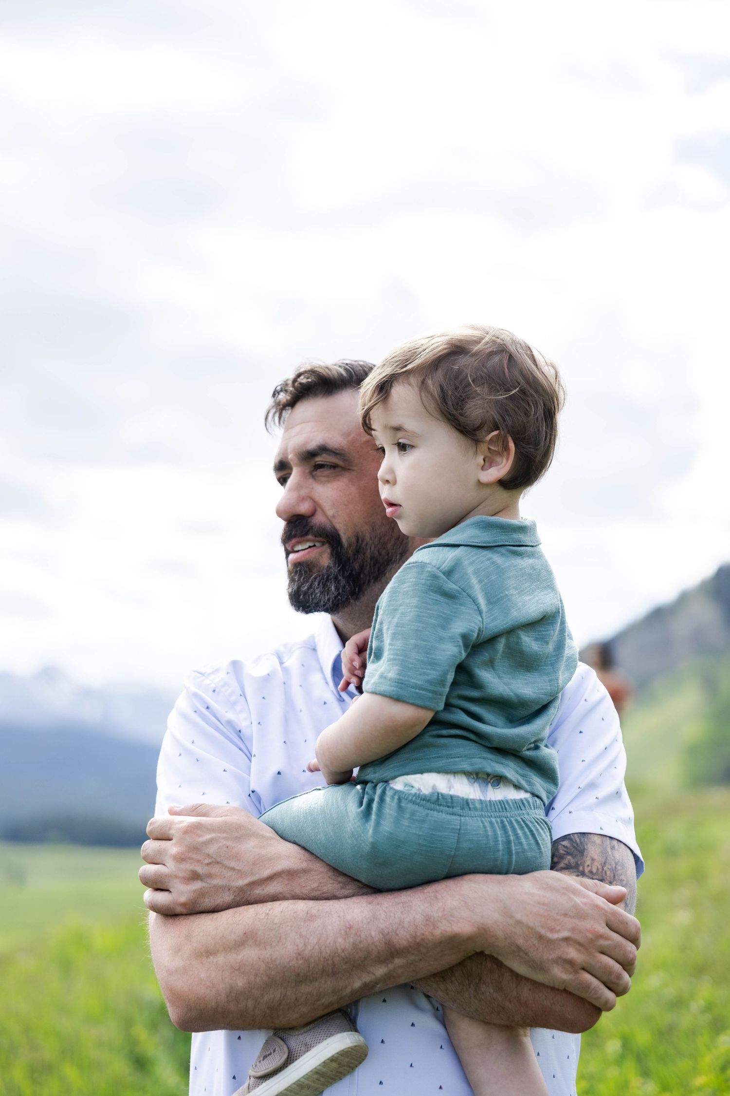 A bearded father holds his young toddler son in a green meadow with mountain scenery in the background.