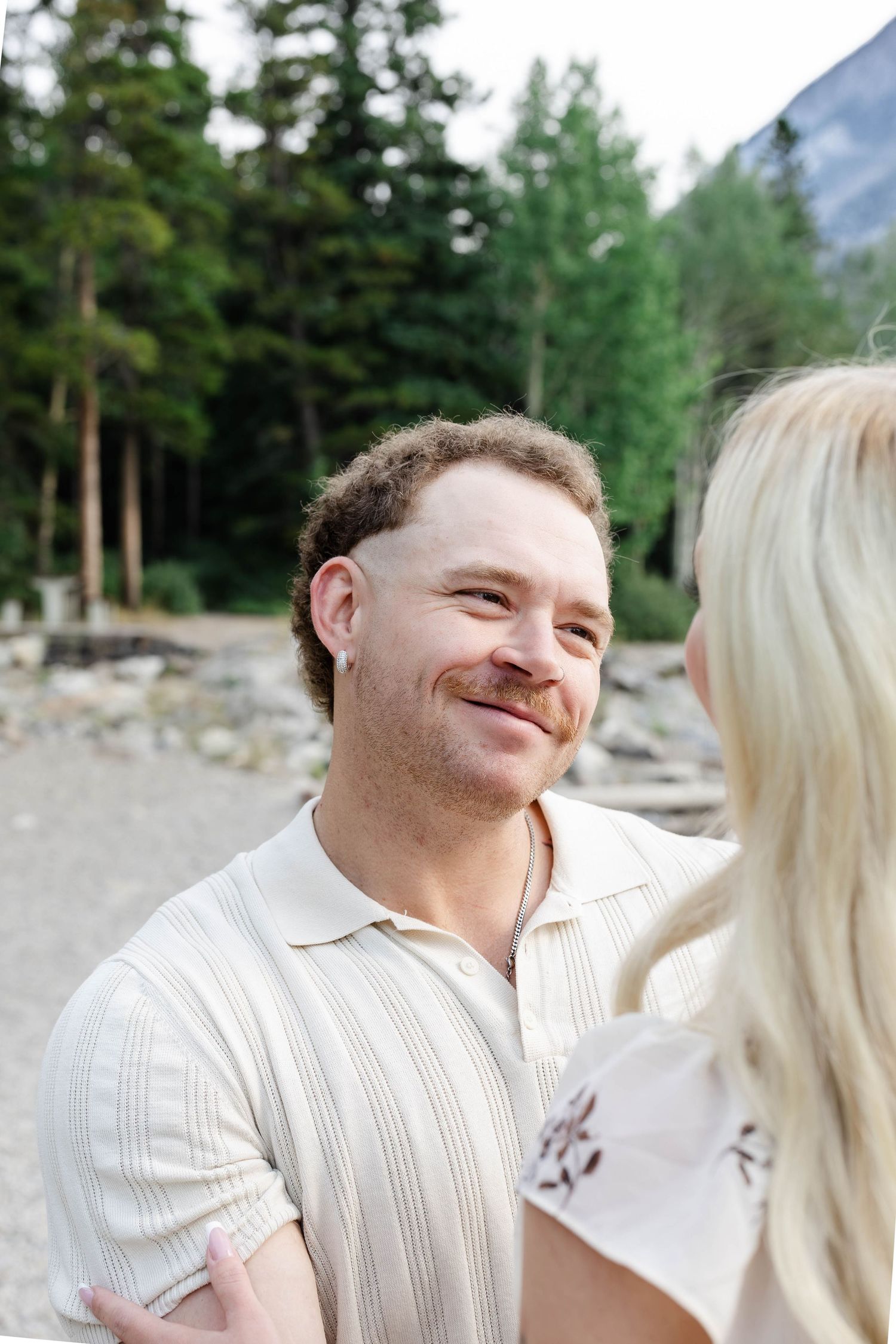 Smiling man with curly hair and ear piercing gazing at blonde woman outdoors near mountain forest.Romantic couple in Minnewanka Lake in Banf