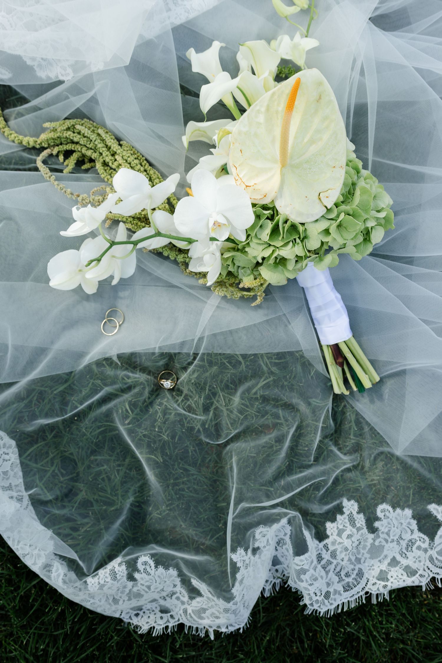 Elegant wedding bouquet with white orchids, calla lilies, and green hydrangeas on lace veil with gold rings.