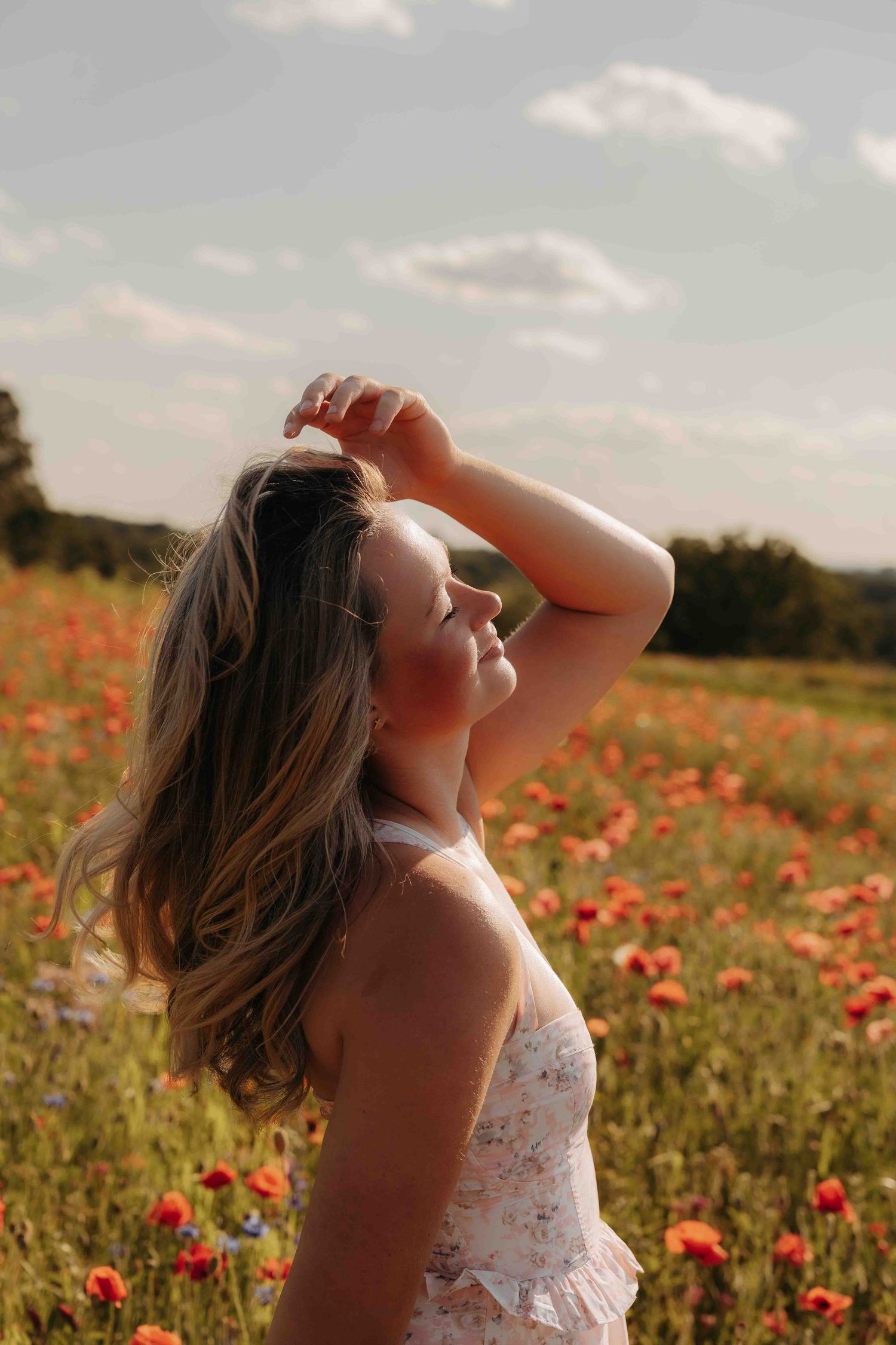 Young woman in floral dress standing in a vibrant red poppy field at sunset.