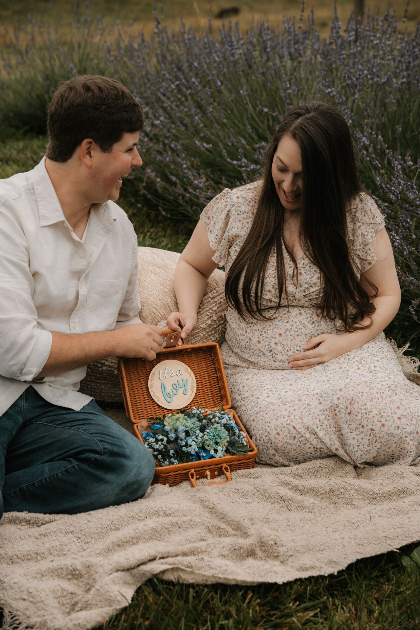 A couple sits in a lavender field revealing a baby gender reveal box with blue flowers during maternity session.