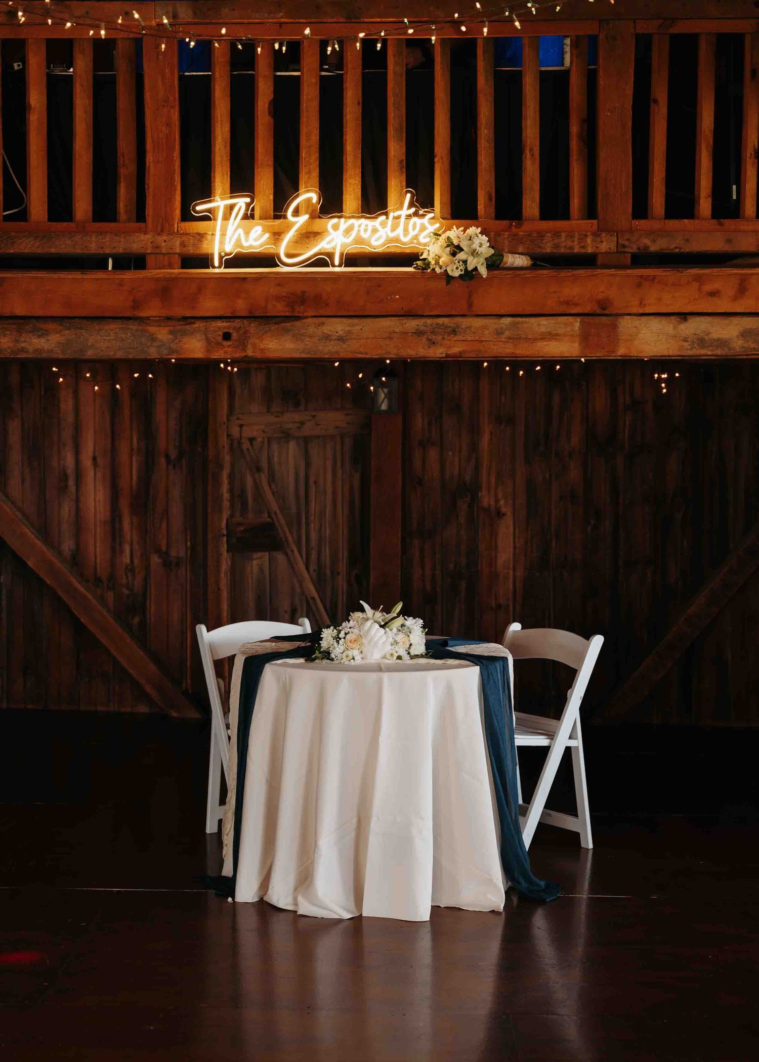 Romantic sweetheart table with navy accents beneath glowing neon The Espositos sign in rustic barn venue.