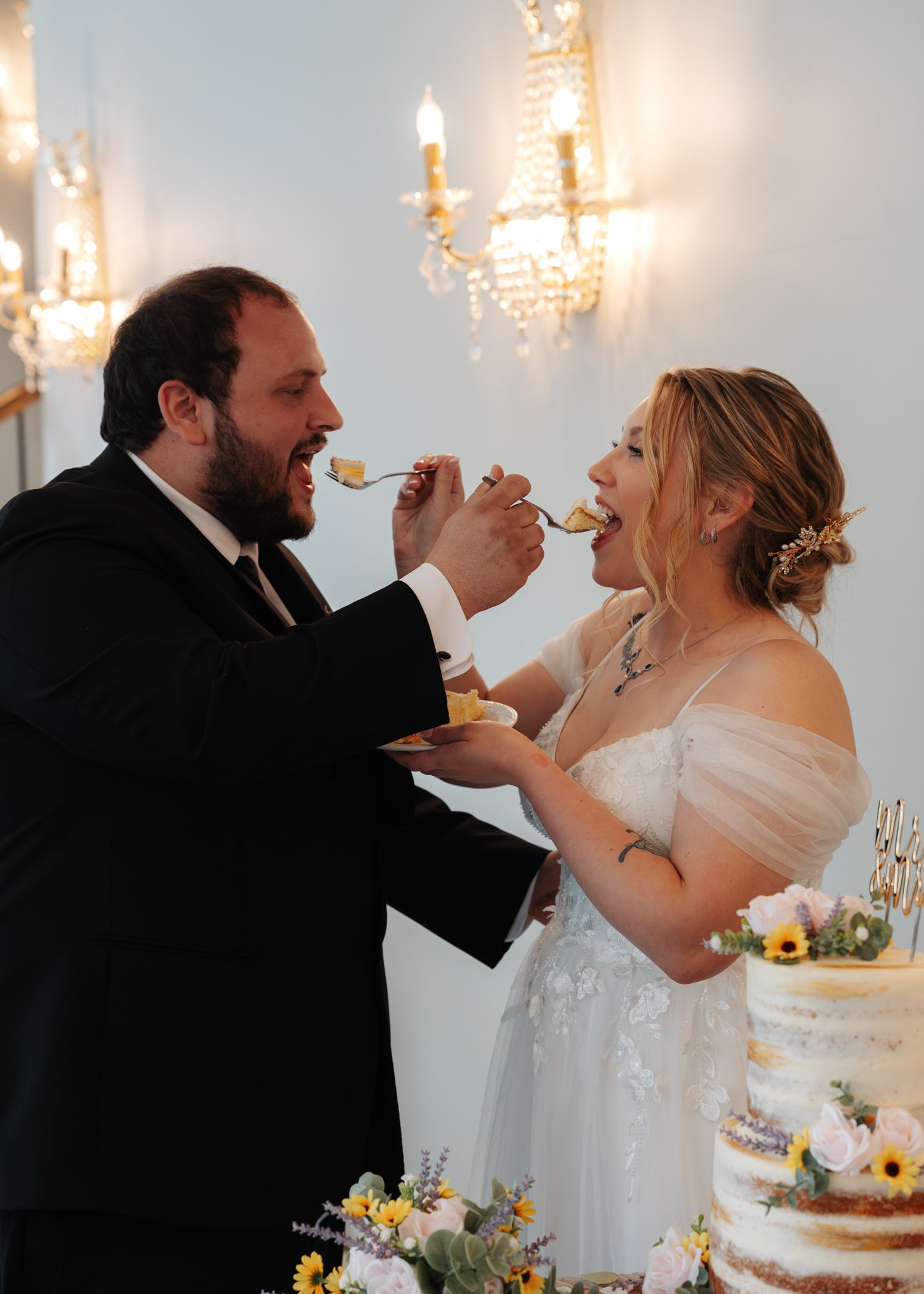 Bride and groom laugh while feeding each other cake beside sunflower-decorated naked wedding cake at reception.