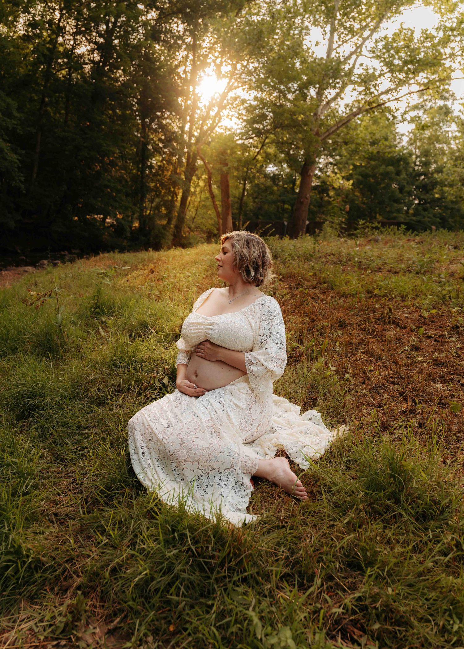 Pregnant woman in white lace dress sitting in sunlit meadow surrounded by green trees at golden hour.