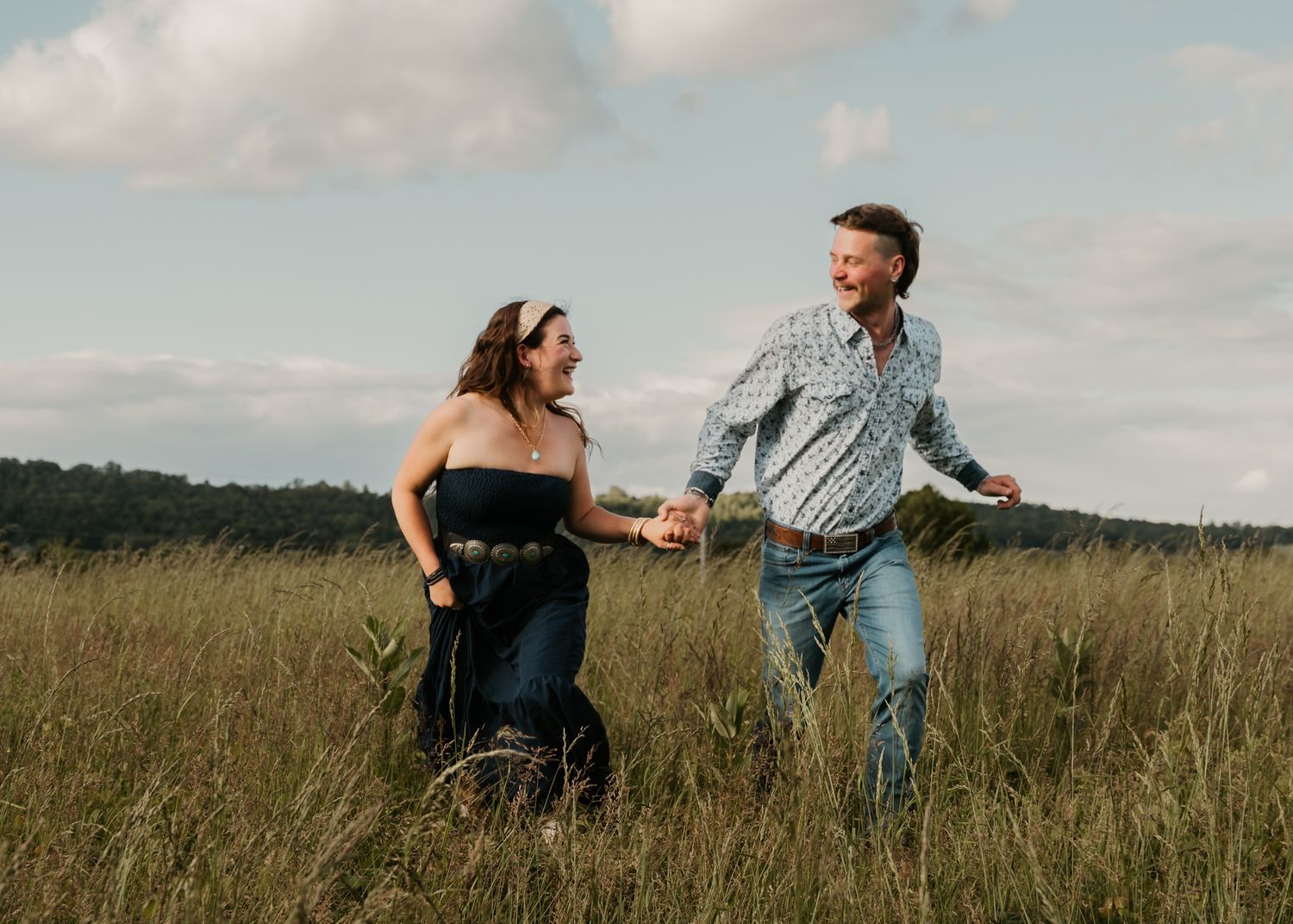 Joyful couple holding hands running through a lush green field under cloudy skies.