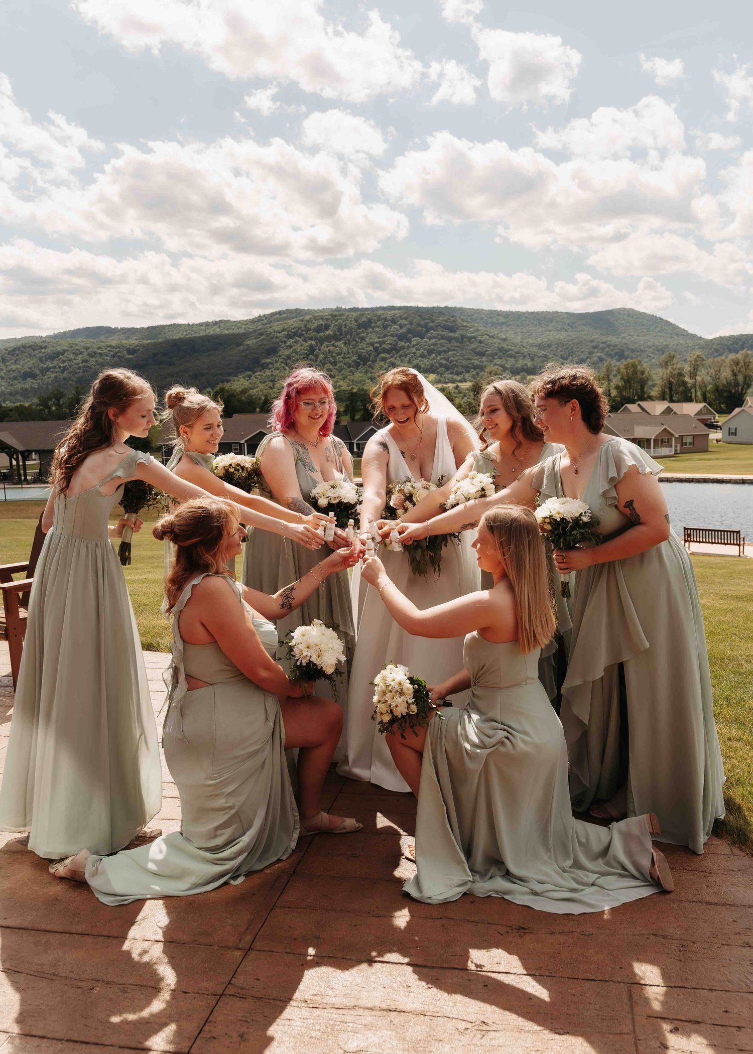 Seven bridesmaids in sage green dresses gather in circle raising bouquets with mountains and clouds in background.