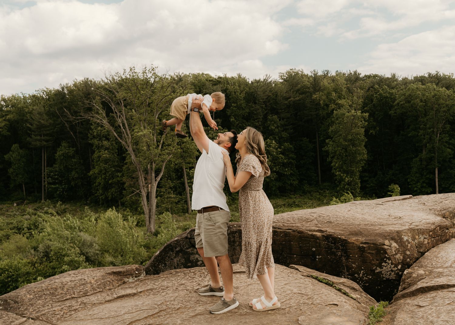 Young couple lifting baby in the air together while standing on large rocks in nature.