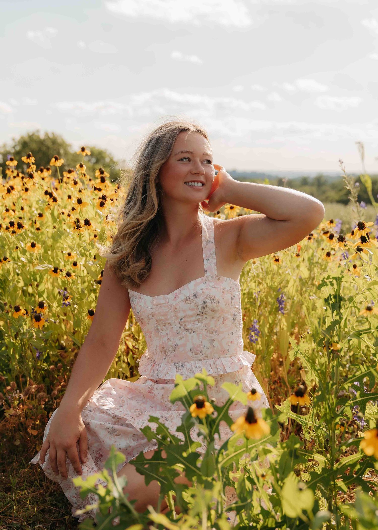 Young woman in a floral dress sitting and smiling in a vibrant wildflower field.