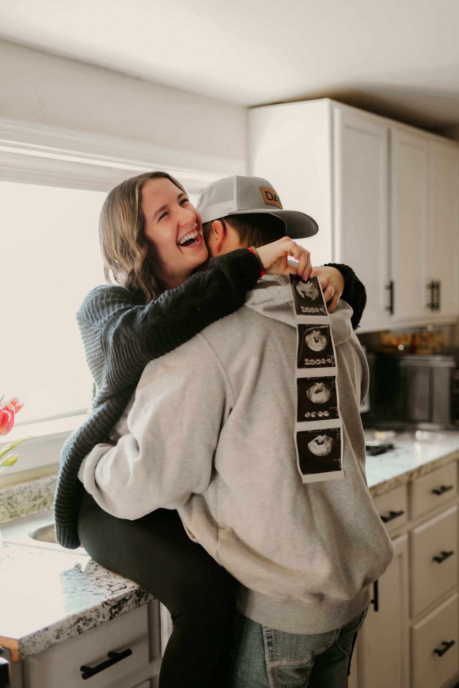 A joyful woman hugs her partner in a kitchen while holding ultrasound photos behind his back for a pregnancy reveal.