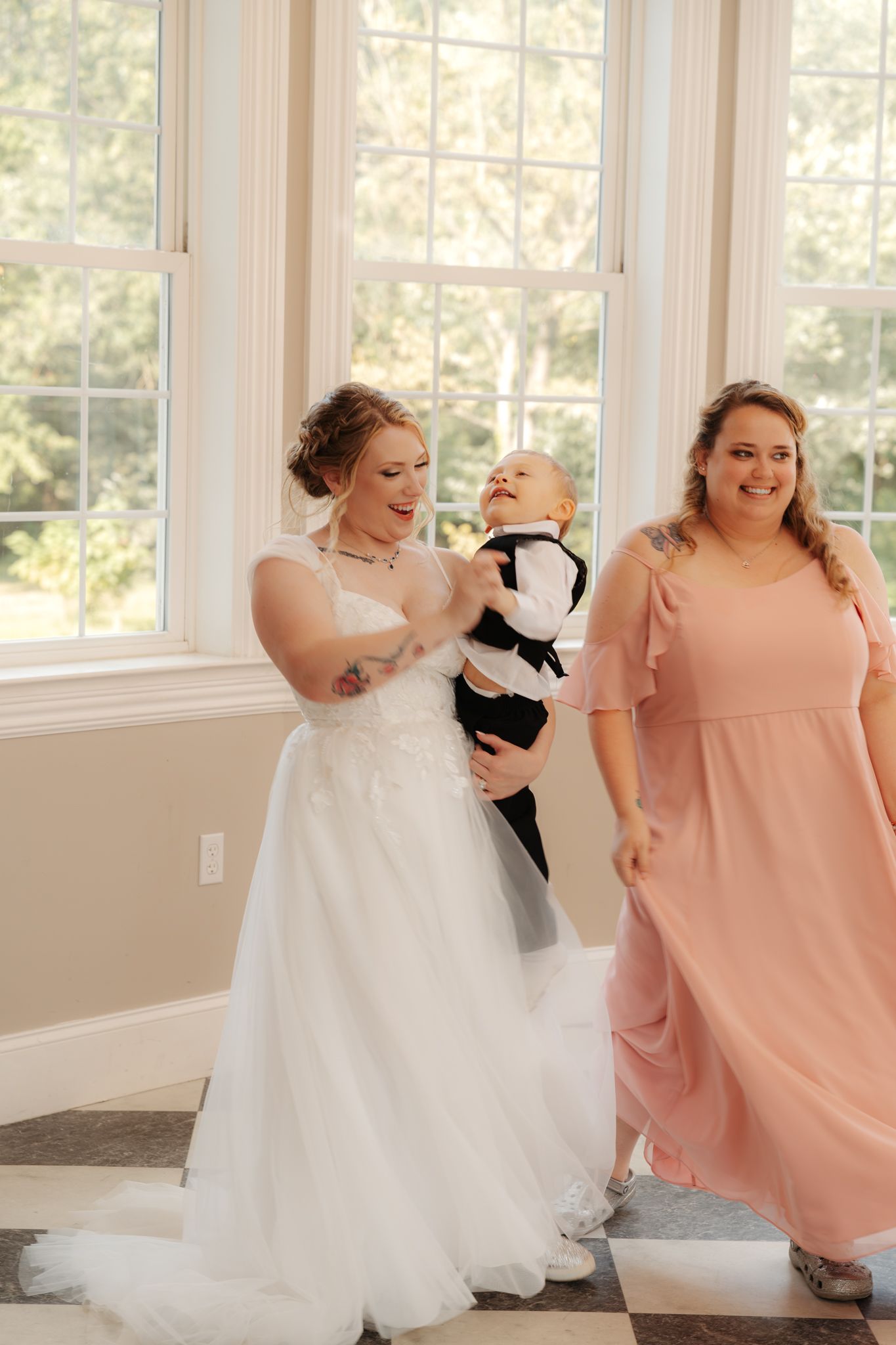 Bride in white gown holding laughing baby in tuxedo with bridesmaid in pink dress.