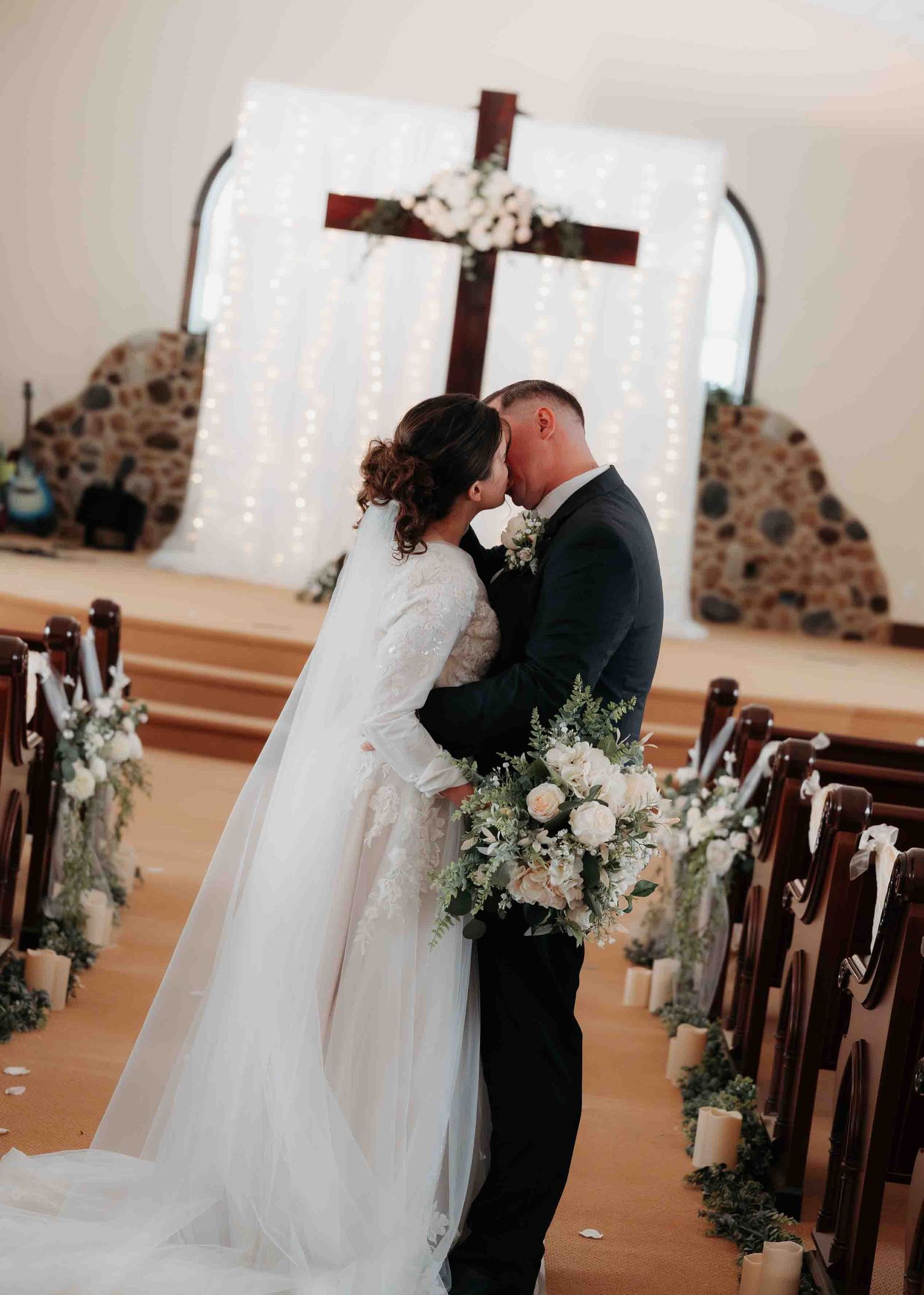 Bride and groom kiss at the altar beneath a wooden cross with fairy light backdrop in church.