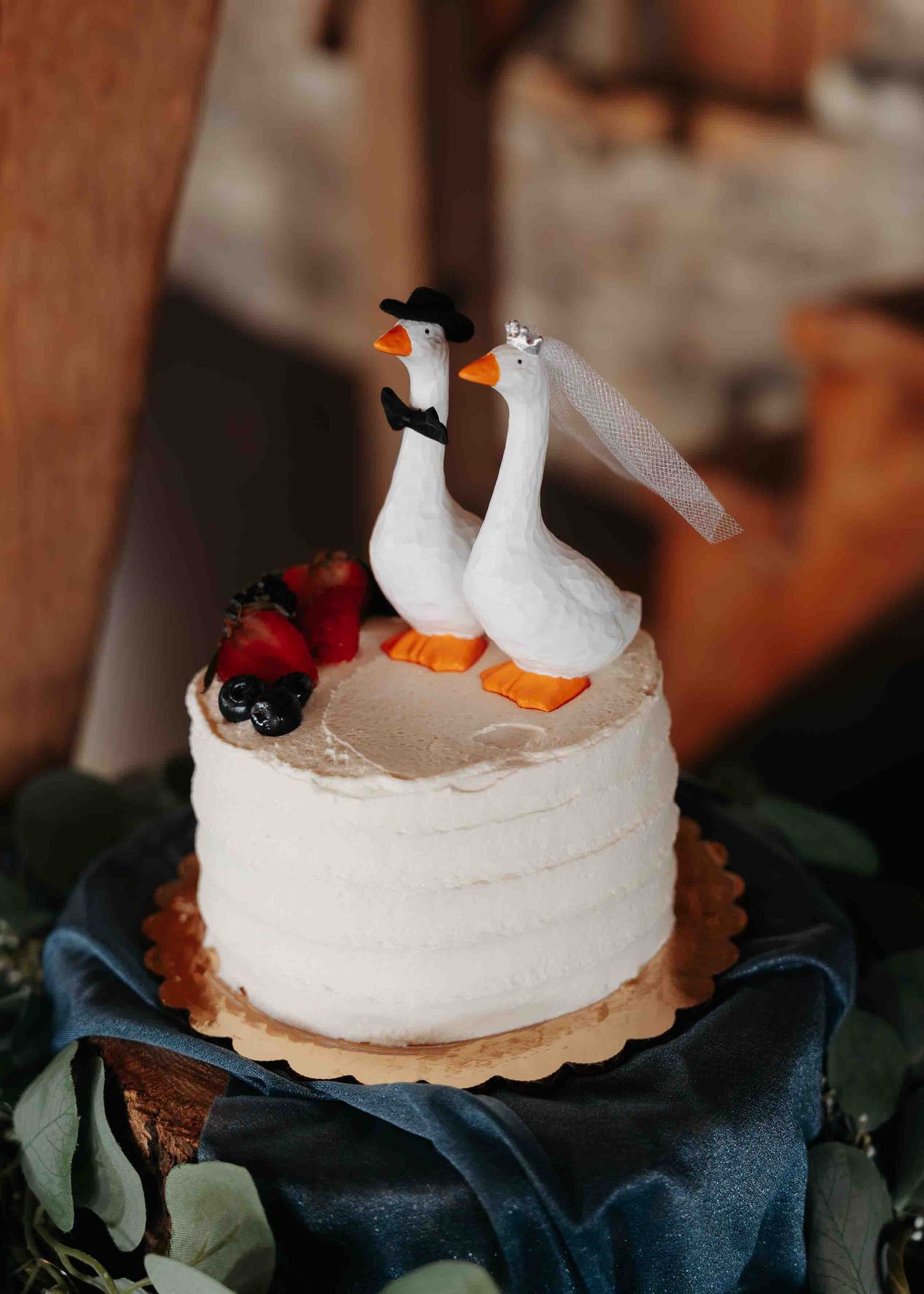 Whimsical wedding cake topped with duck bride and groom figurines decorated with fresh berries and greenery.