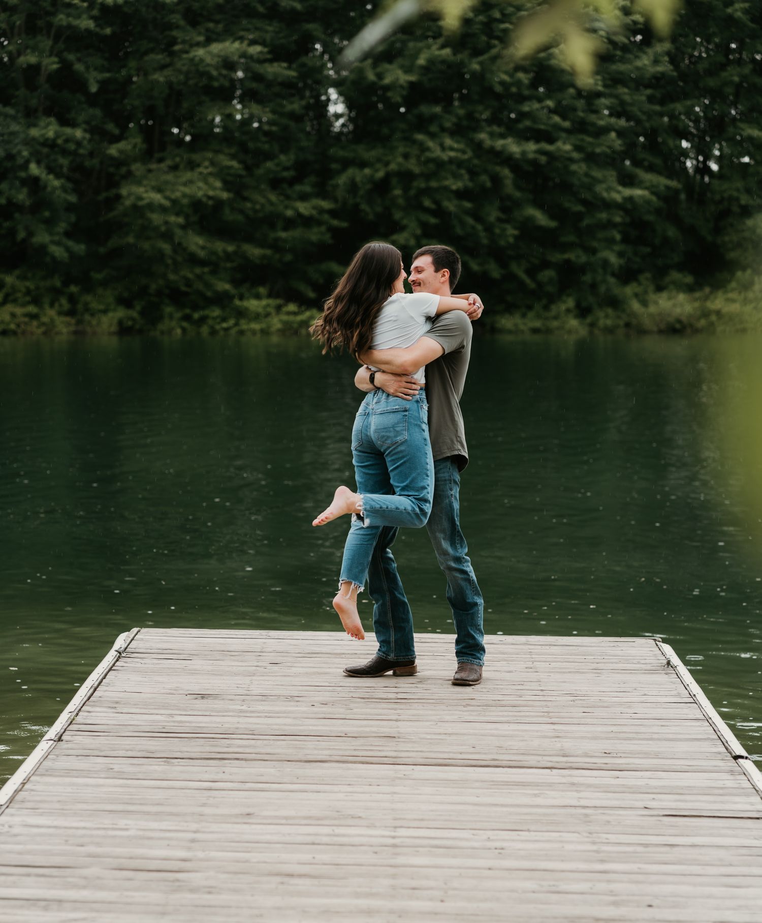 Couple embracing and spinning on a wooden dock overlooking a calm green lake.