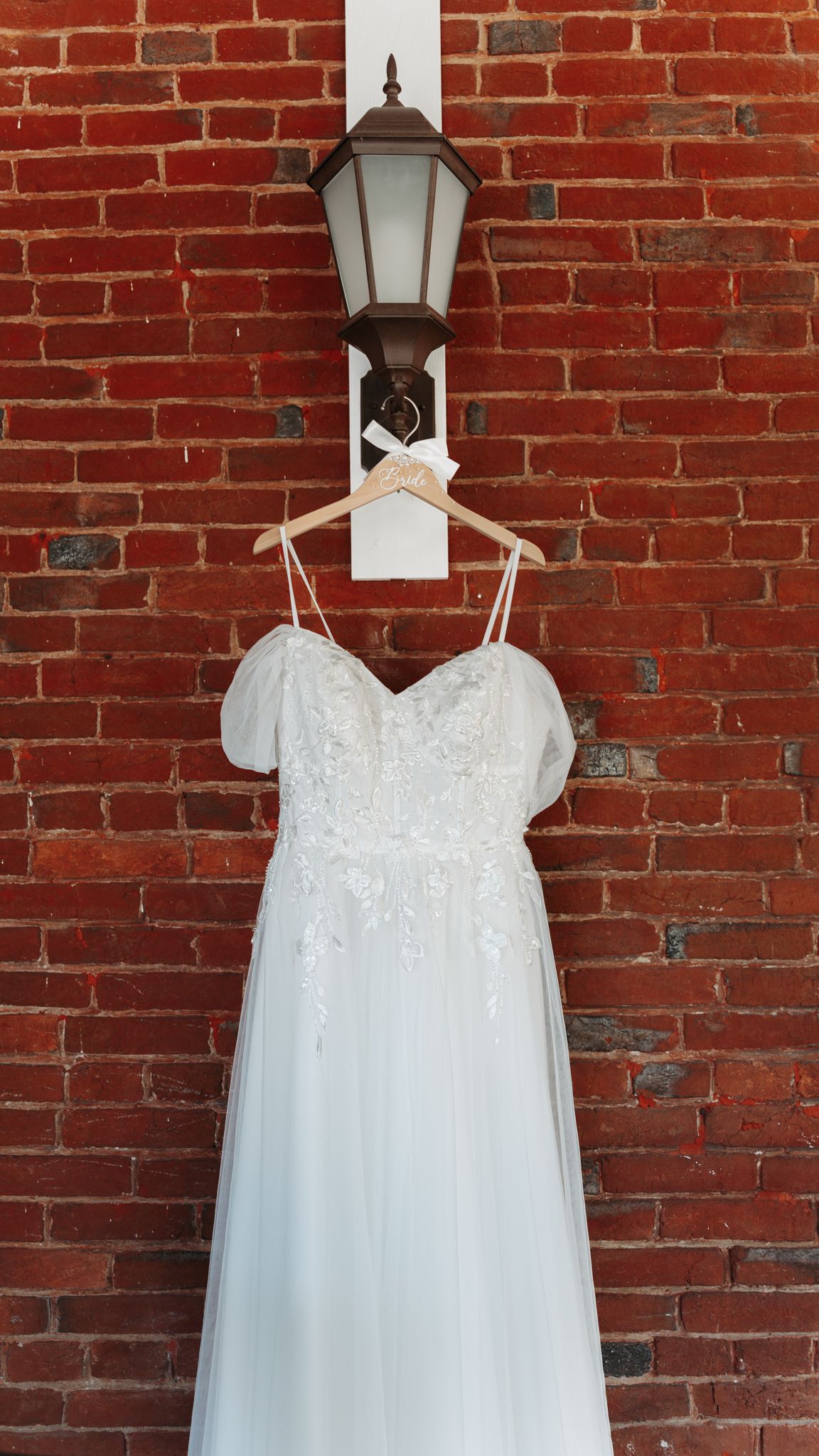 Elegant white lace wedding dress hanging on a wooden hanger against a red brick wall.