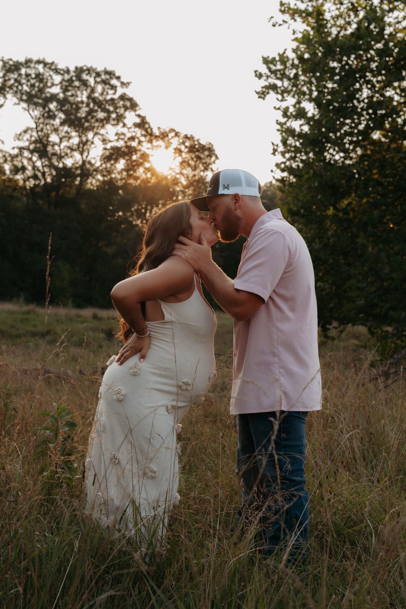 Couple sharing a kiss at golden hour in tall grass field at sunset.