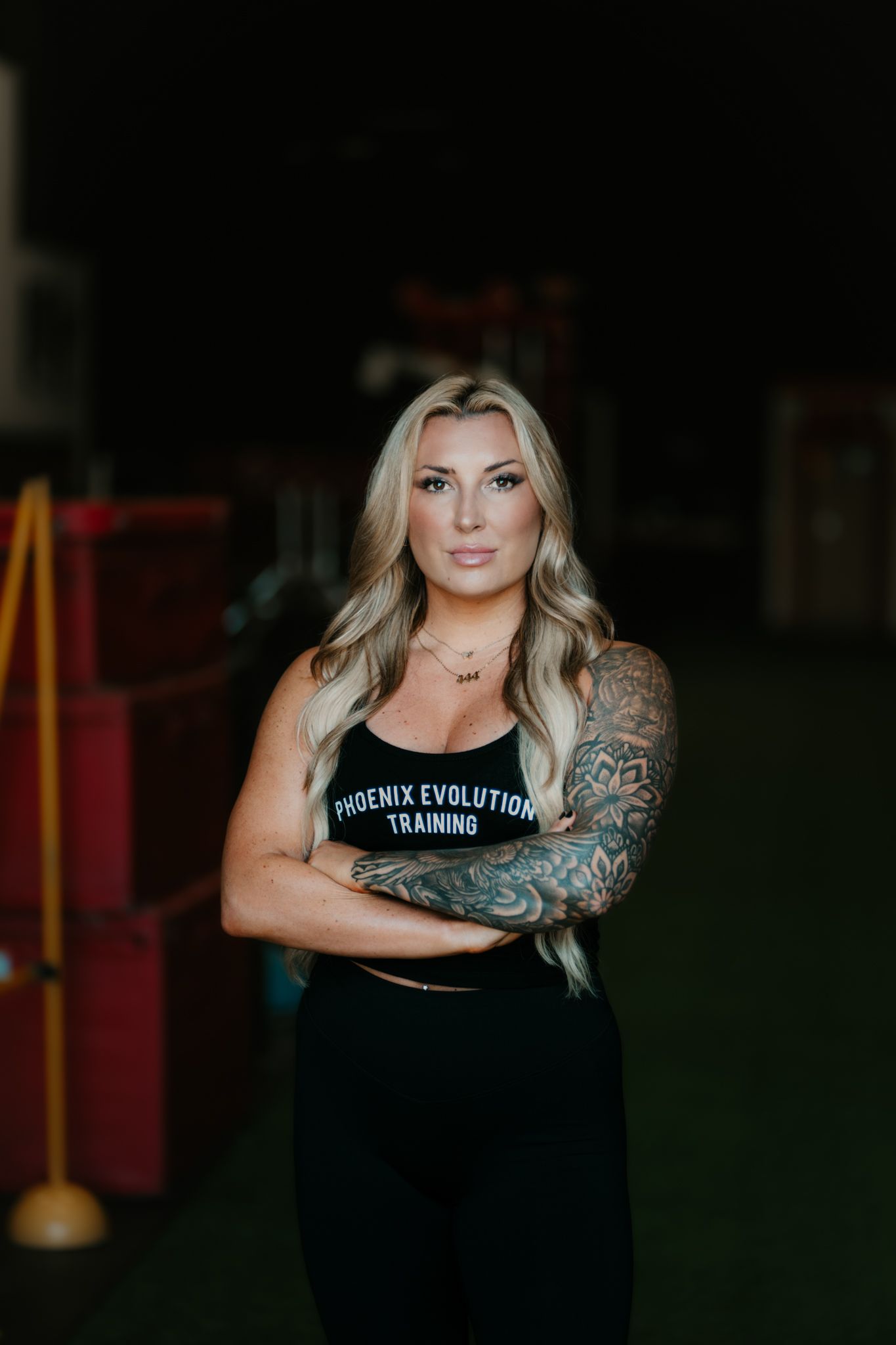 Tattooed blonde woman in Phoenix Evolution Training top poses with arms crossed in gym.