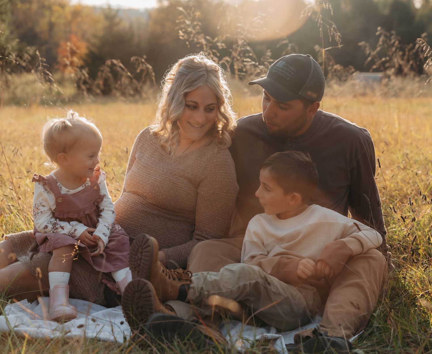 Family of four sitting together on a blanket in a sunlit autumn field, smiling warmly.
