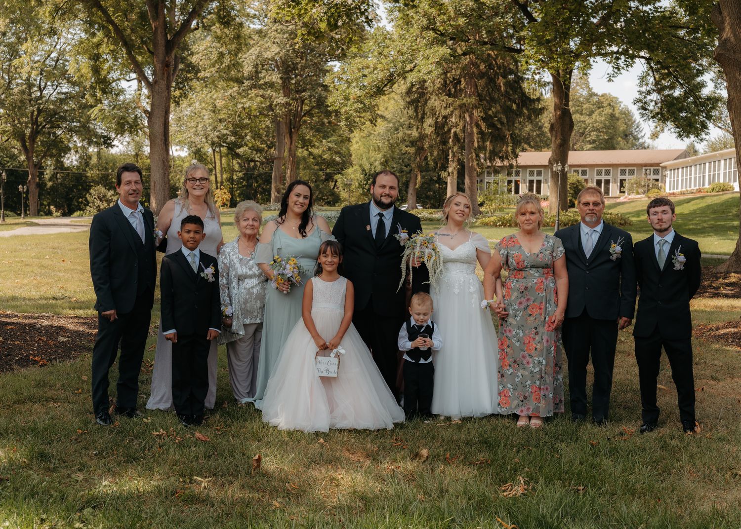 Wedding party group portrait with bride, groom, bridesmaids, groomsmen, and children on green lawn.