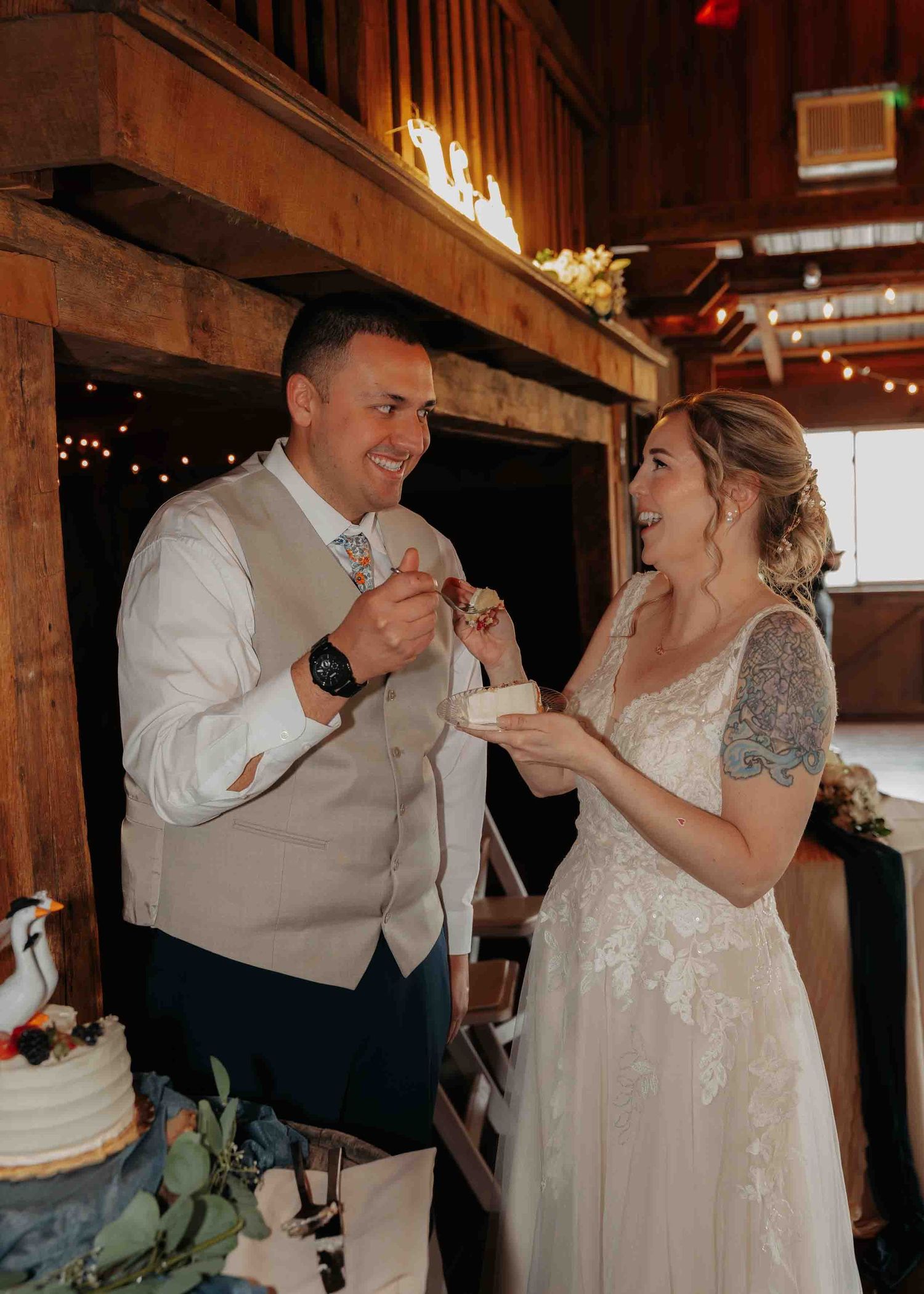 Laughing newlyweds enjoy cake cutting moment together in rustic barn wedding reception with string lights.