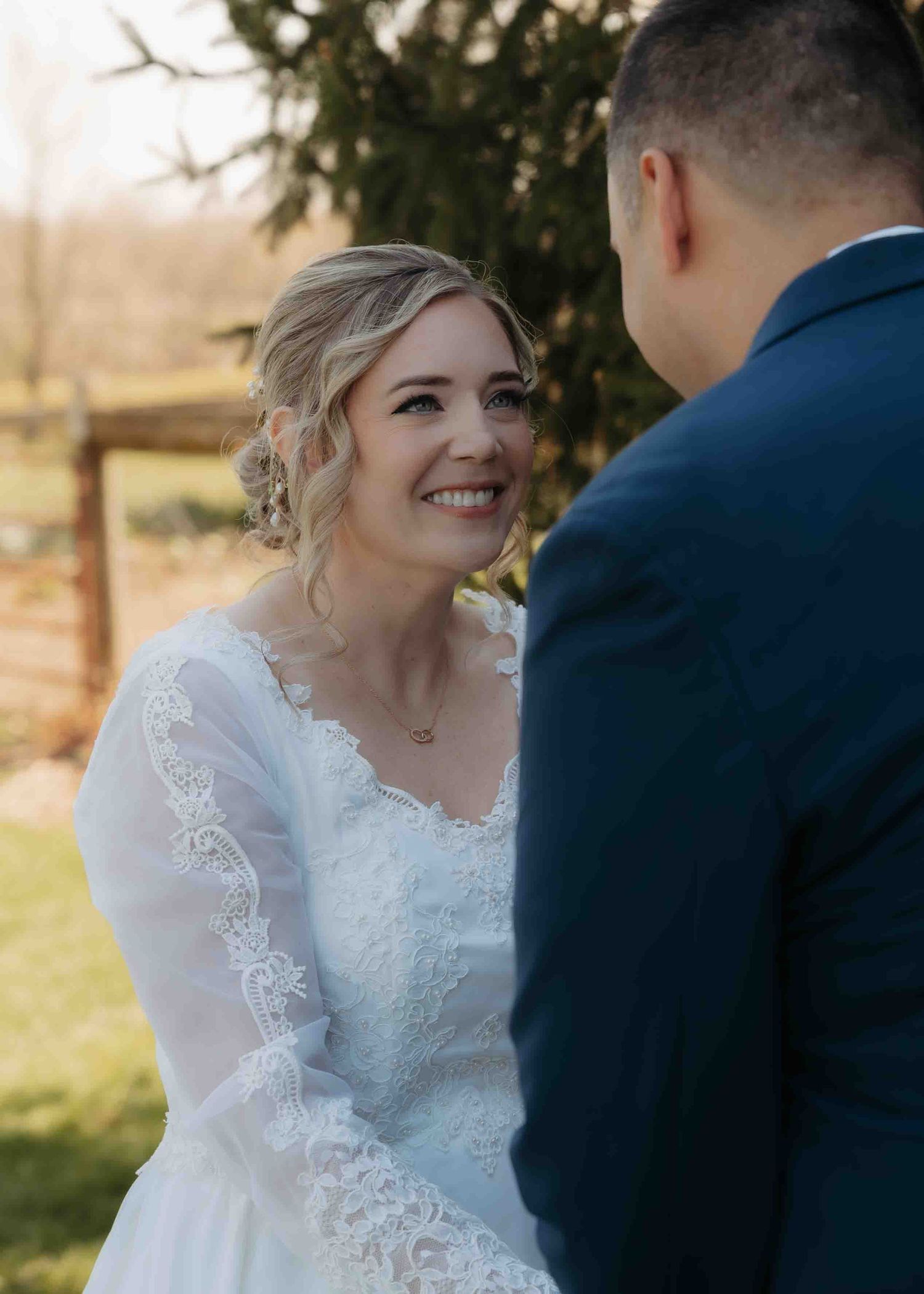 Glowing bride in lace wedding gown smiles warmly at groom during outdoor ceremony.