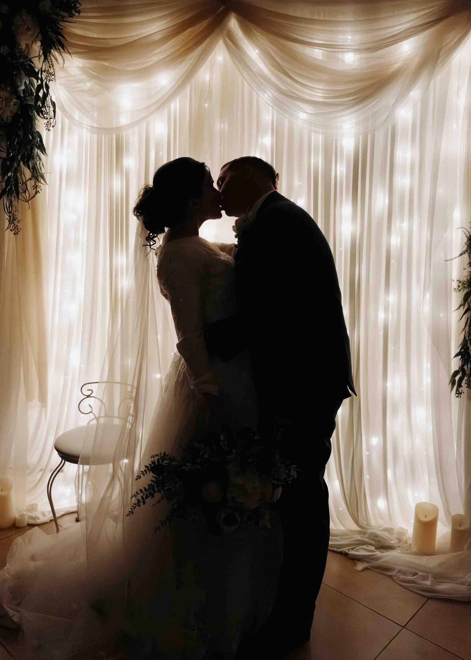 Romantic silhouette of bride and groom kissing in front of a glowing fairy light curtain backdrop.