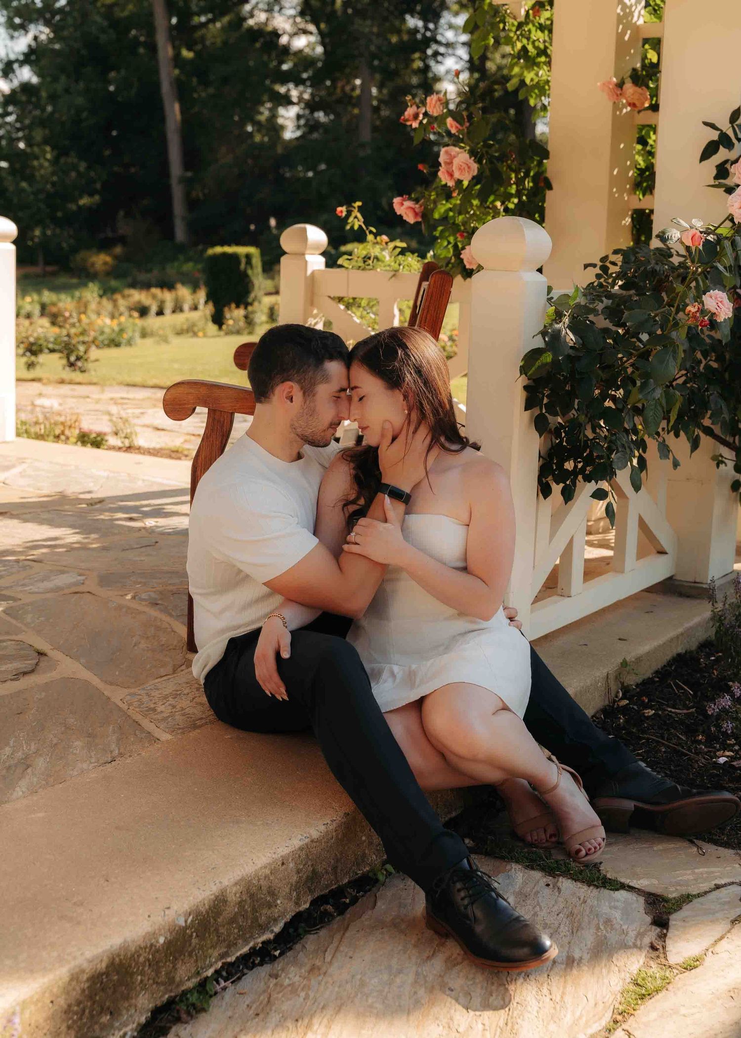 Couple sits close together on garden steps surrounded by blooming roses at golden hour.