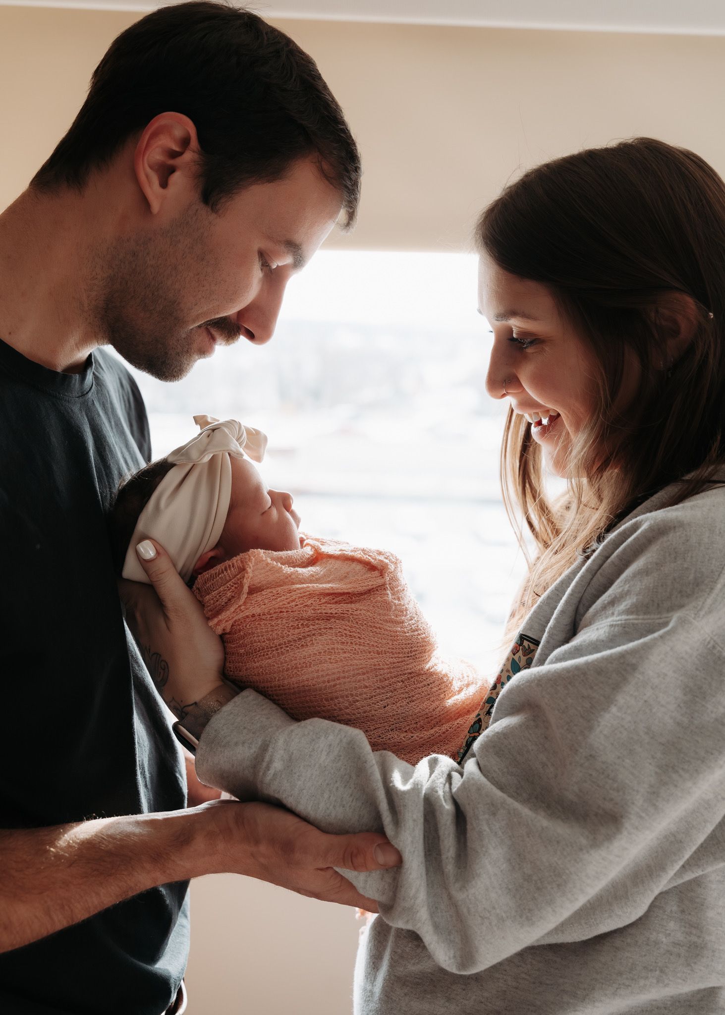 Happy new parents smiling at their swaddled newborn baby in a bright room.