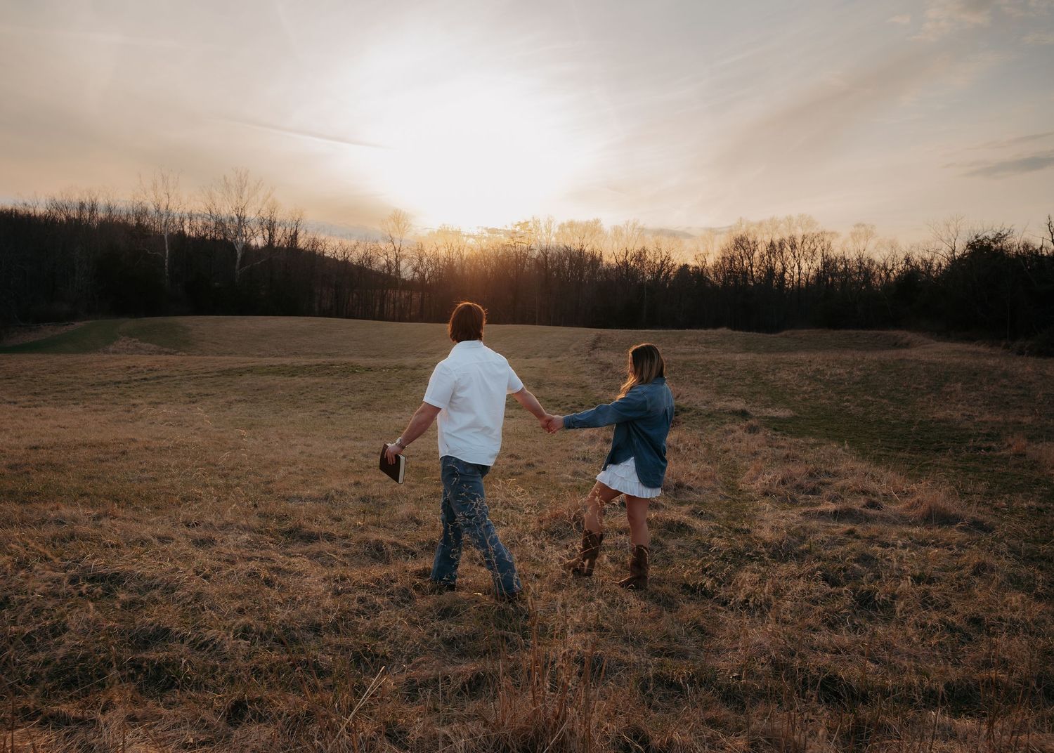 Couple holding hands walking toward sunset across a golden field in a romantic countryside setting.
