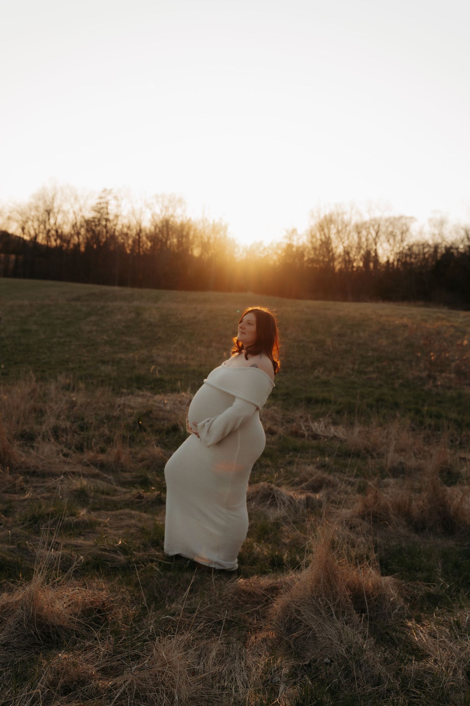 Pregnant woman in flowing white dress standing in golden field at sunset during maternity shoot.