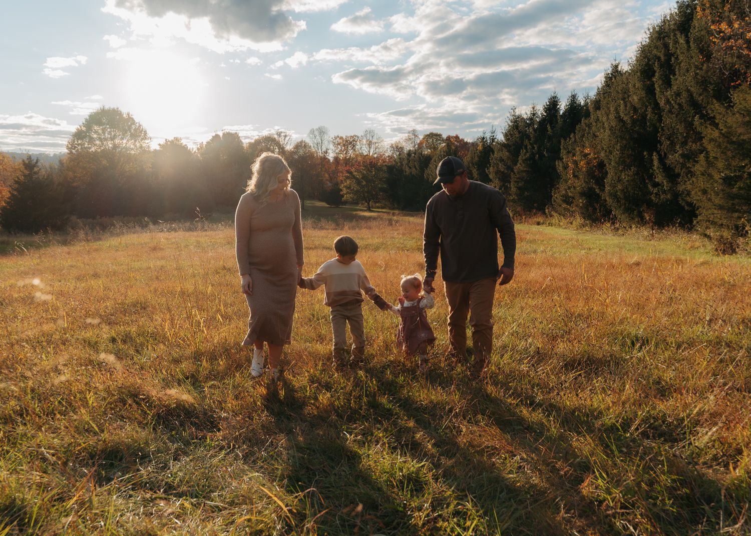 Family of four walks toward sunset in a golden meadow surrounded by autumn trees.