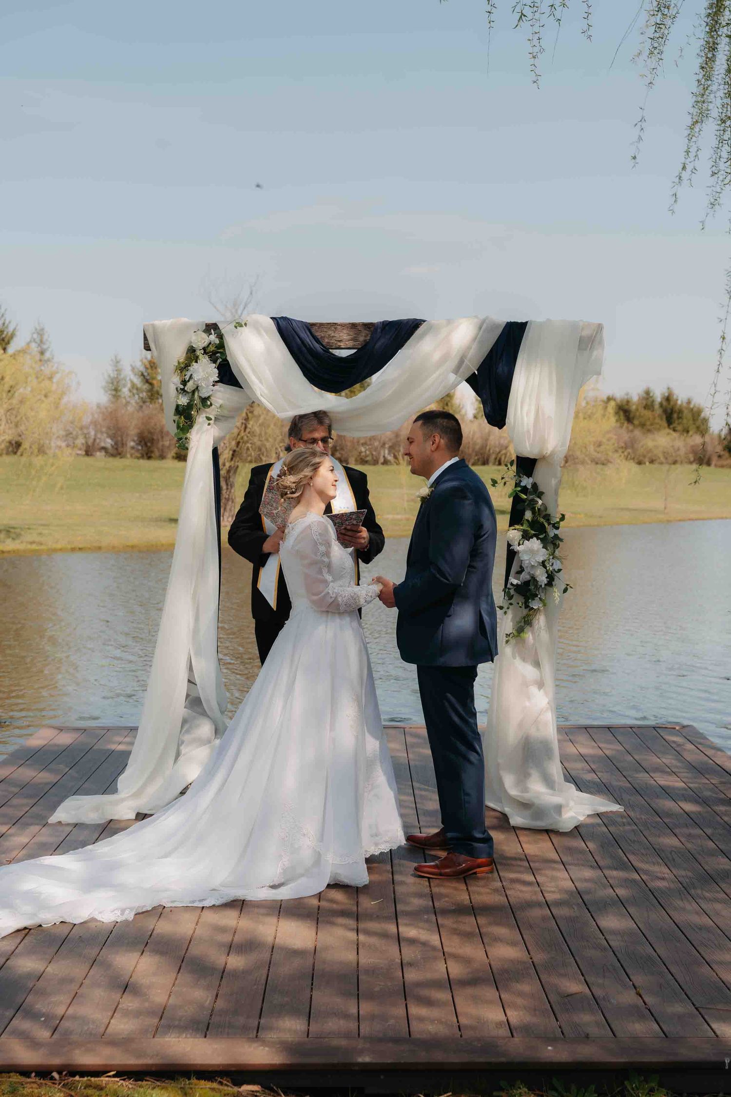Couple exchanges vows at outdoor waterfront altar adorned with white draping and florals.