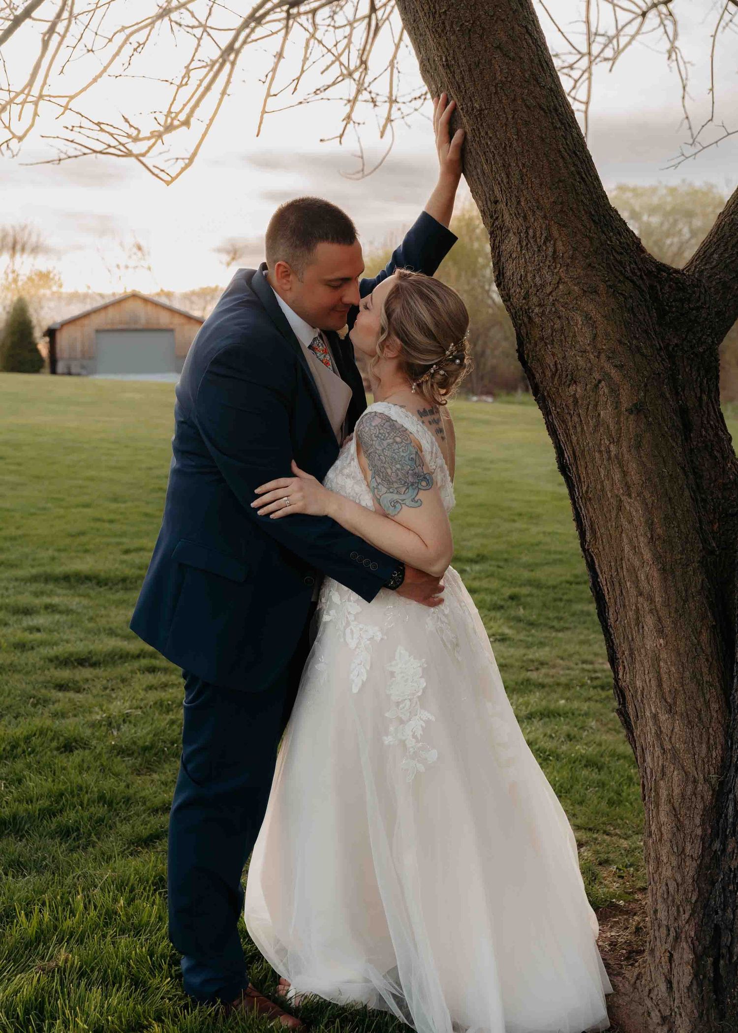 Bride and groom sharing romantic kiss by tree at golden hour outdoor wedding.