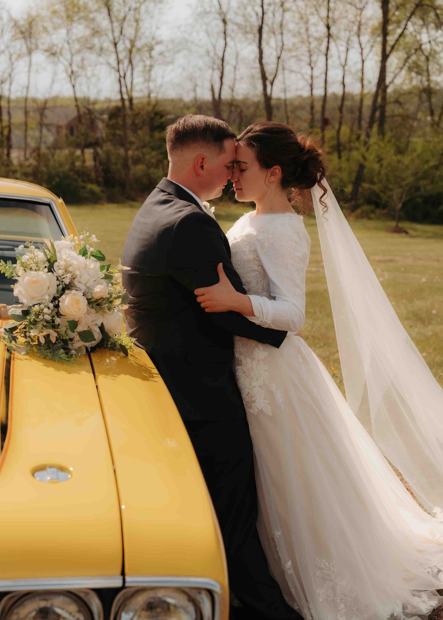 Newlyweds share an intimate moment leaning against a classic yellow car outdoors.