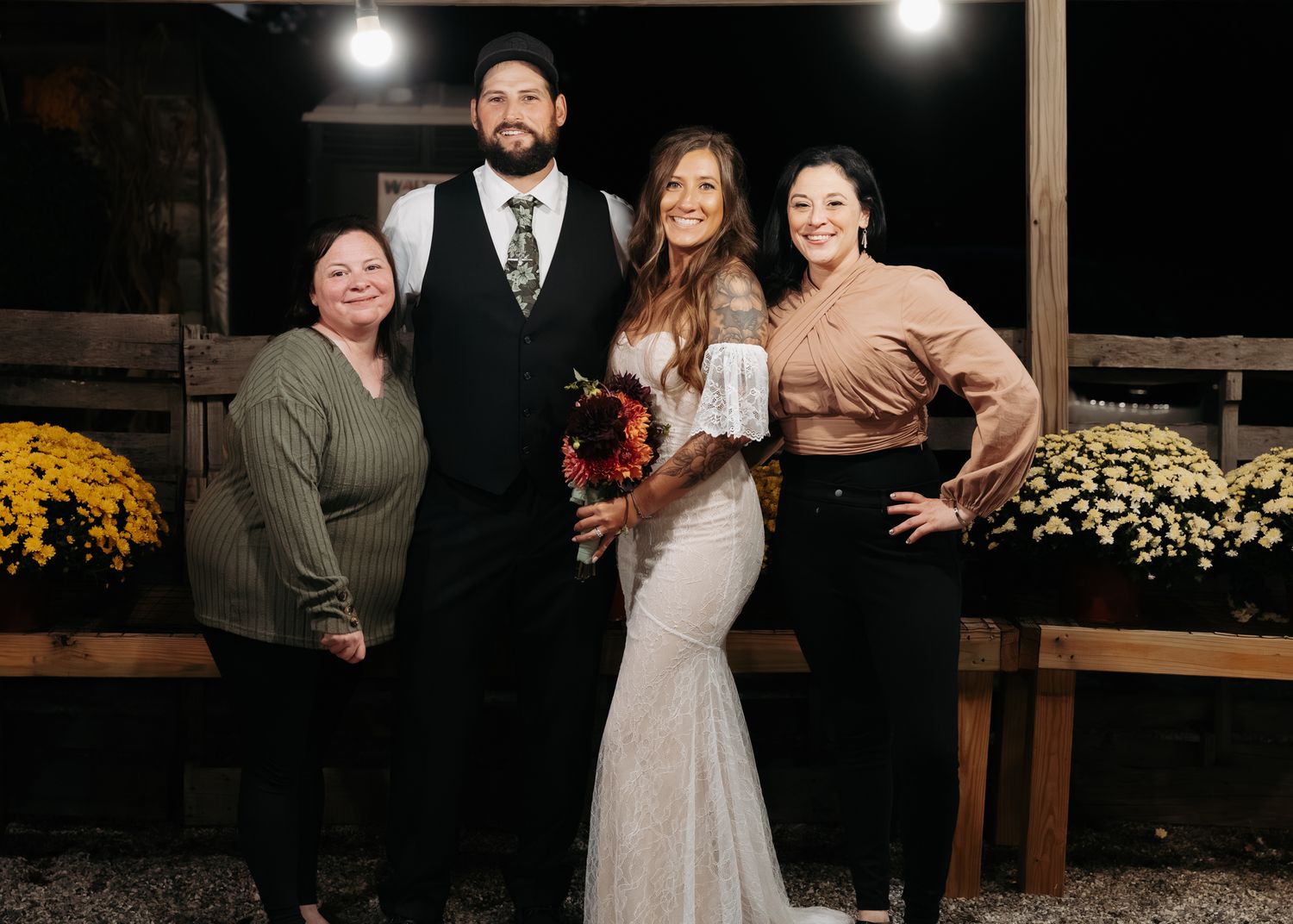 A wedding group photo featuring a bearded groom in a vest and his bride in a lace gown, flanked by two smiling women.