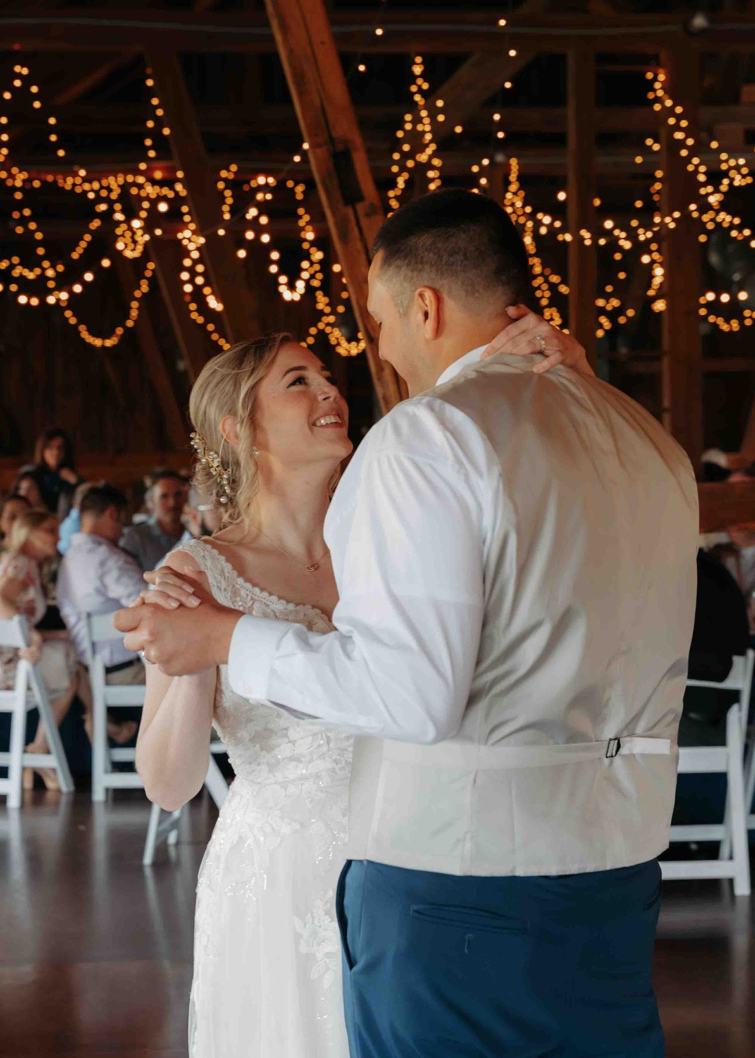 Bride and groom share emotional first dance under warm string lights in a rustic barn reception venue.