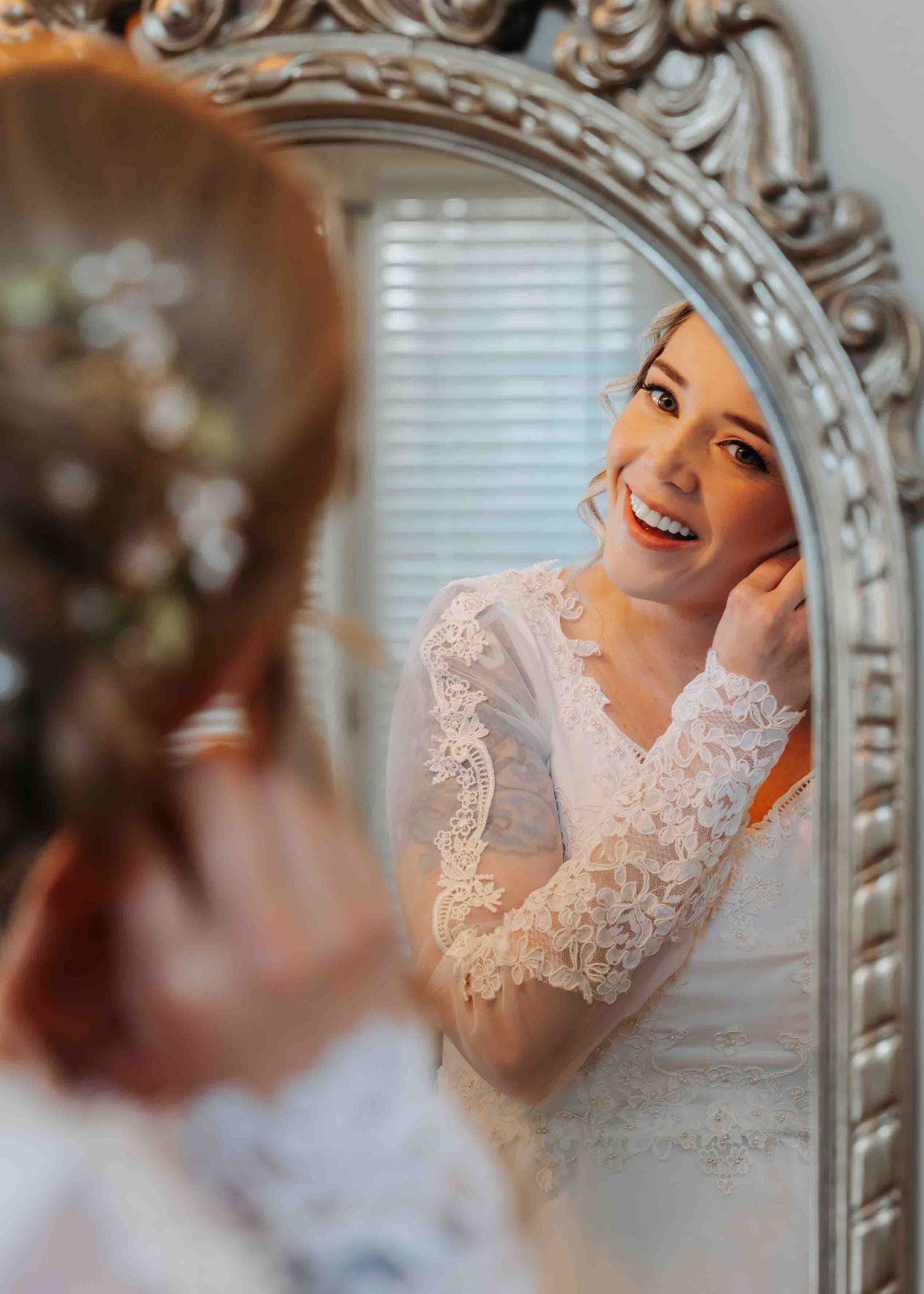 Smiling bride in lace wedding dress adjusts earring while looking in ornate mirror.