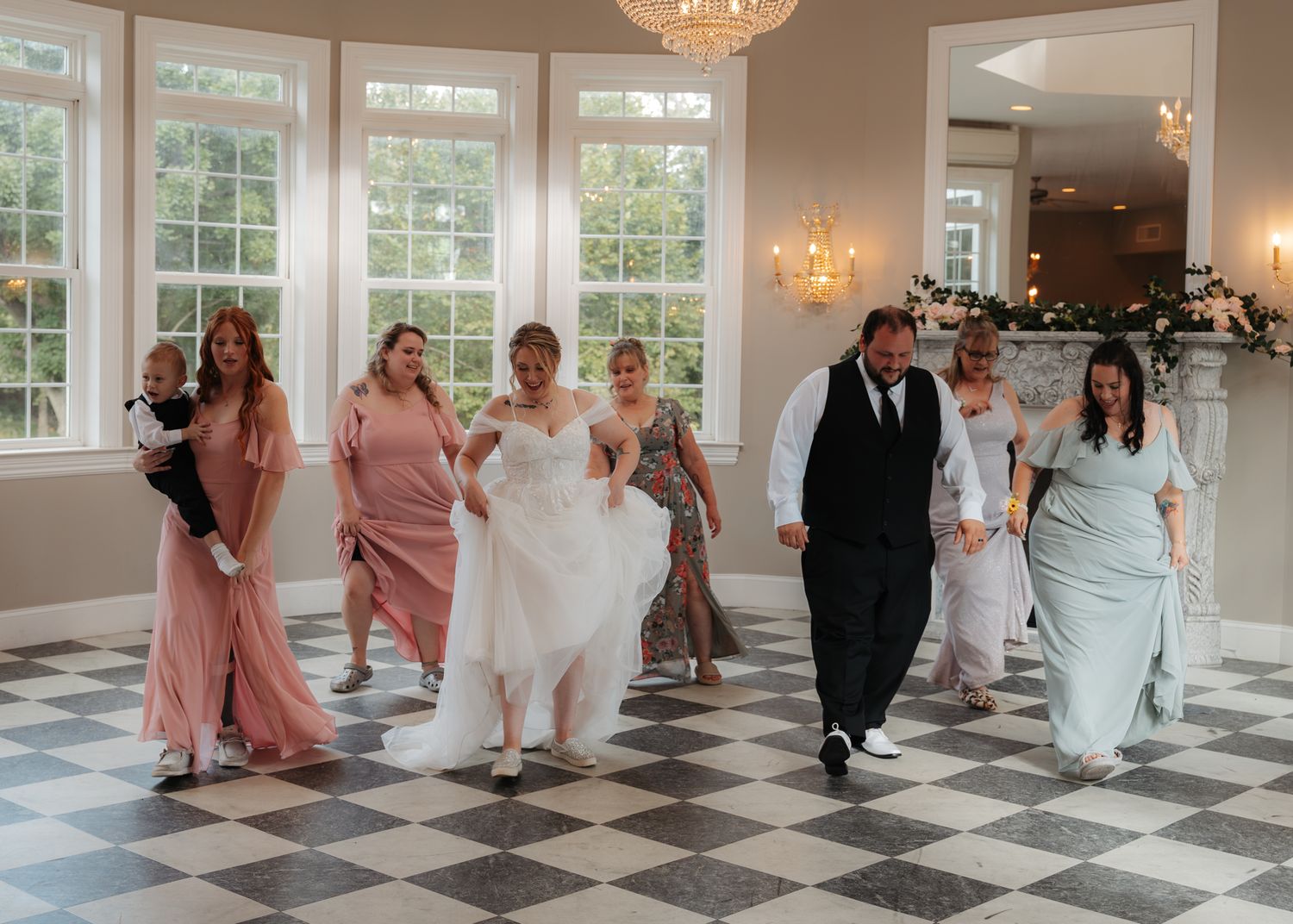 Wedding party walking through elegant ballroom with checkered floors and floral decorations.