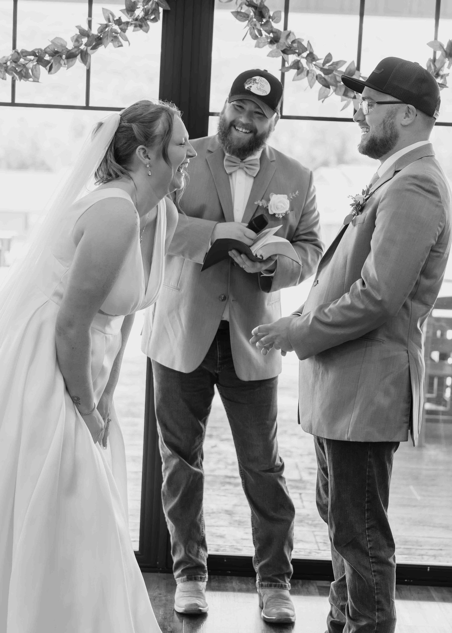 Black and white photo of bride and bearded groom exchanging vows with officiant at indoor wedding ceremony.
